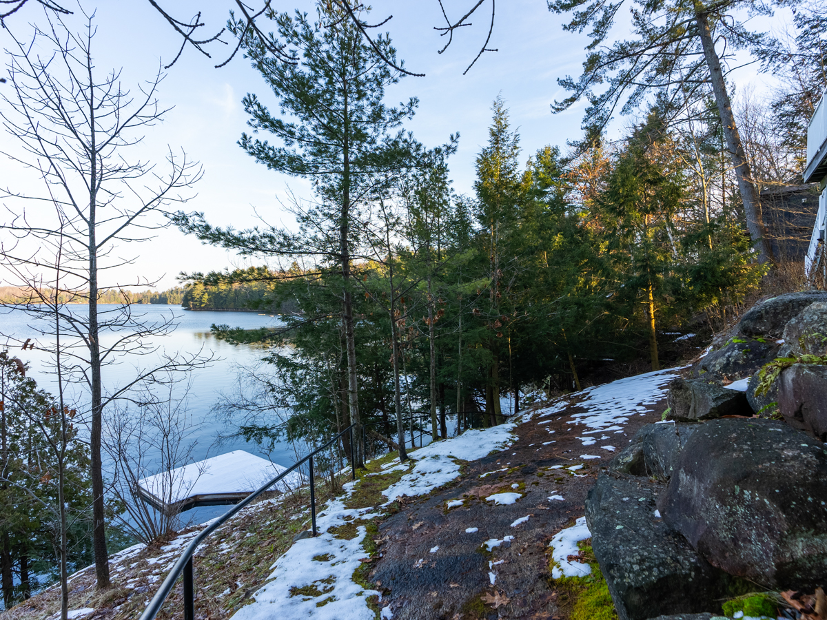 A wide path extends along a sloping, rocky waterfront property. A metal railing traces the edge of the path toward the water. A big snow-covered dock extends into the blue lake down below..