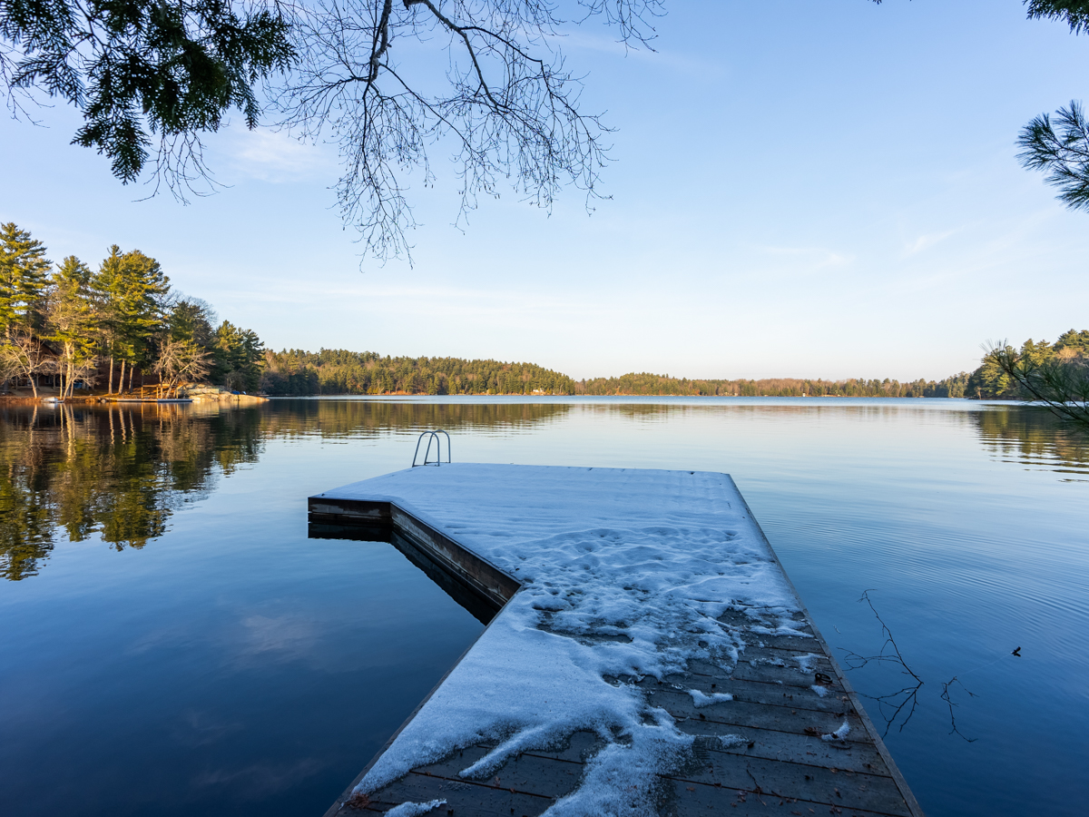 A big snow-covered dock extends out into a still blue lake. Across the water, the shore is lined with green trees.