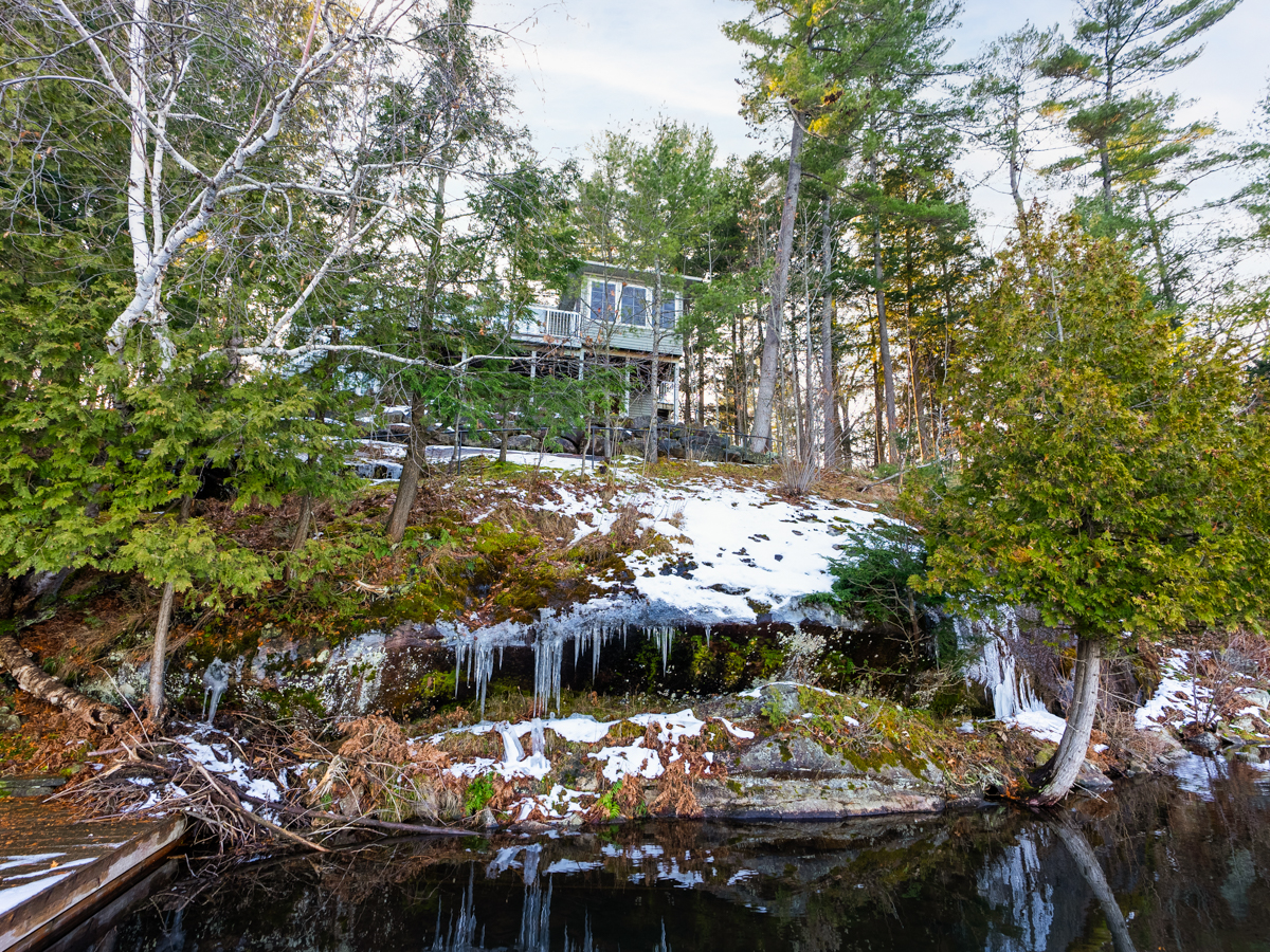 The shore of a lake, with natural rock and trees creating a big hill. Icicles hang off the edge of a big rock. A large cottage sits at the top of the hill, and down at the shoreline, a big dock extends into the water.