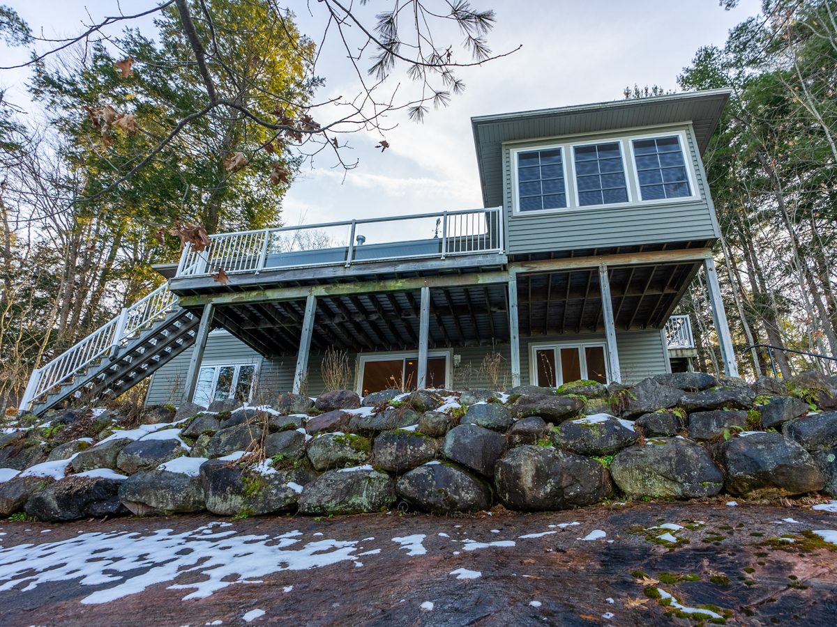 Looking up from a rocky natural area at a big cottage that sits at the top of the slope. The cottage is two storeys, and it has lots of windows and a big deck facing the water.