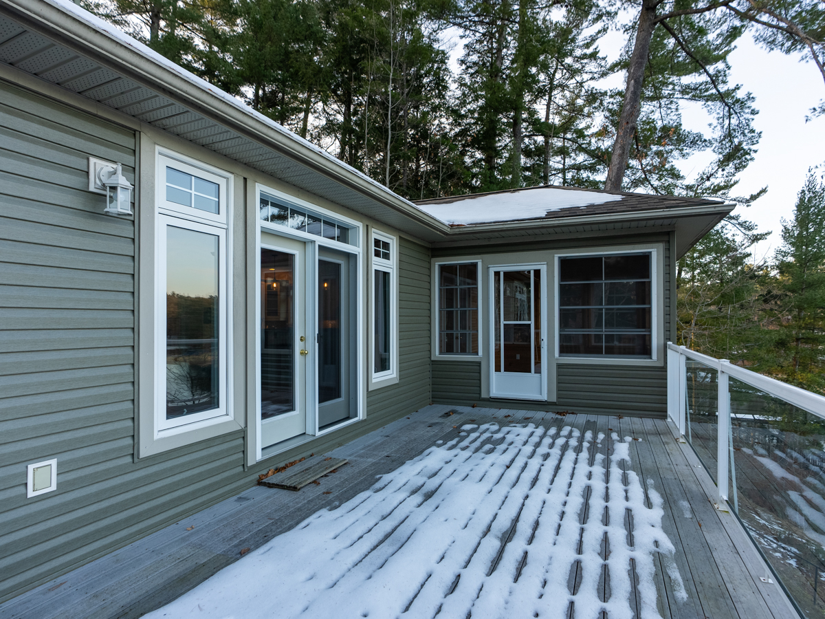 A wide snow-covered deck extends off the back of a big cottage with lots of windows. Two sets of doors lead inside, on adjacent walls.