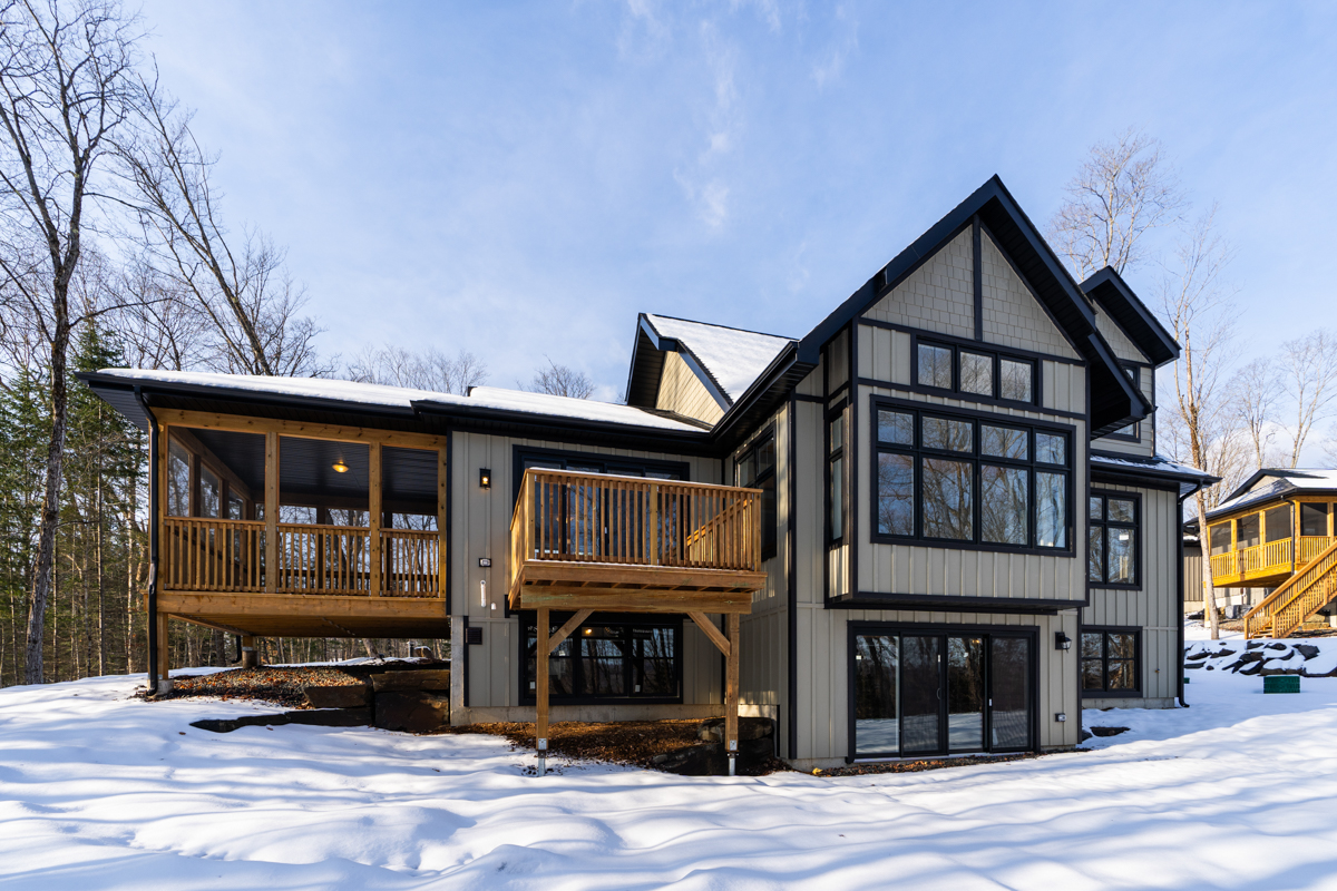 The back of a large two-story home in a rural area. The house has big windows with black trim, a raised deck, and a sunroom that extends off the back of the house. There is snow on the ground.