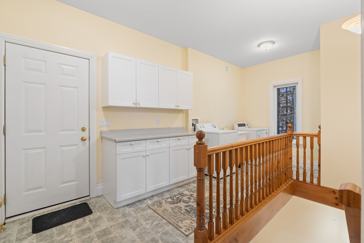 An entrance area with a door and tiled flooring. A narrow space stretches out beside it with a counter that has upper and lower cabinets. A washing machine and dryer sit side by side at the end of the space. There is a wood railing in front of the laundry area, and on the other side of it, stairs lead down.