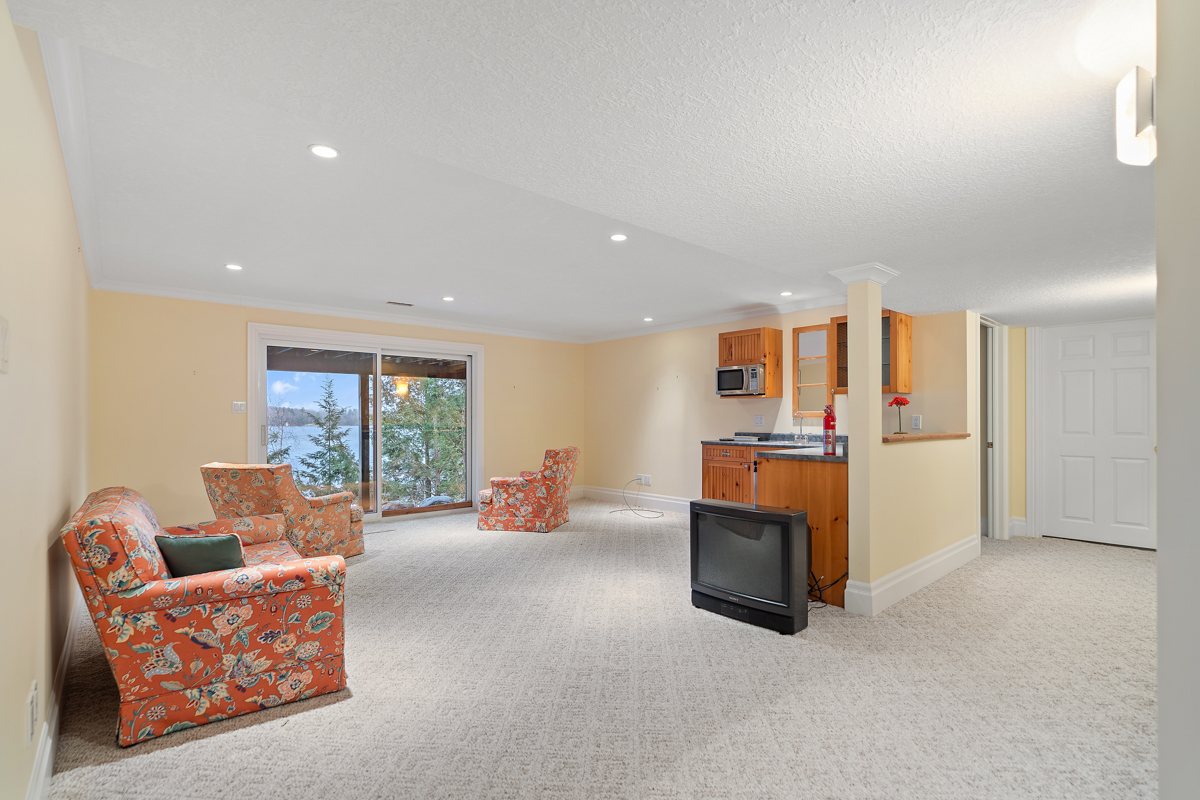 A bright basement living space with yellow walls, carpet flooring, and three pink chairs. Double glass doors walk out to an outdoor space. The room has a small kitchen corner with a sink.