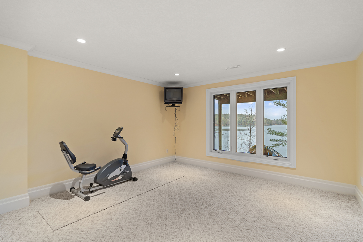 A big empty room with yellow walls, carpet flooring, and a big window. There is an exercise machine against one wall and a small television mounted in the corner.