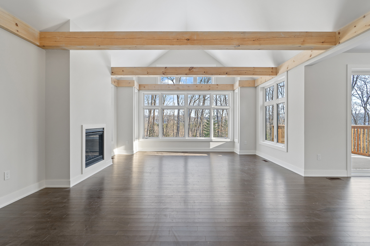 A bright, empty space with dark hardwood floors, white walls, a fireplace, and exposed beams of light wood spanning a white vaulted ceiling. Large windows look out to a forested area with bare trees.