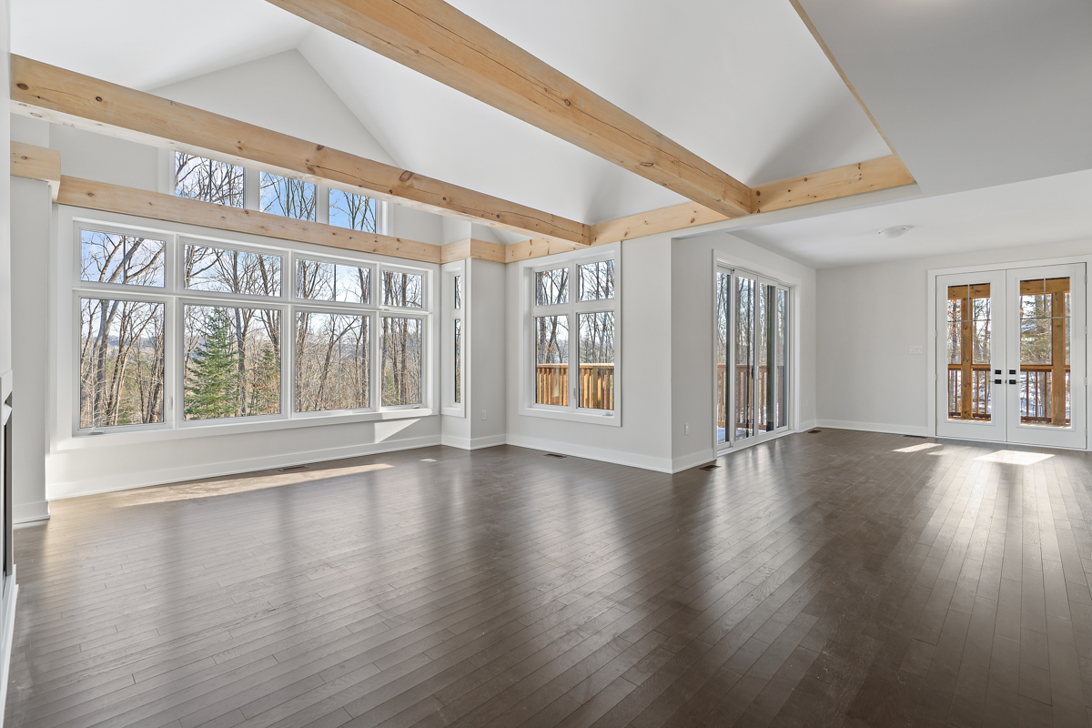 A bright, empty space with dark hardwood floors, white walls, and exposed beams of light wood spanning a white vaulted ceiling. Large windows look out to a forested area with bare trees.