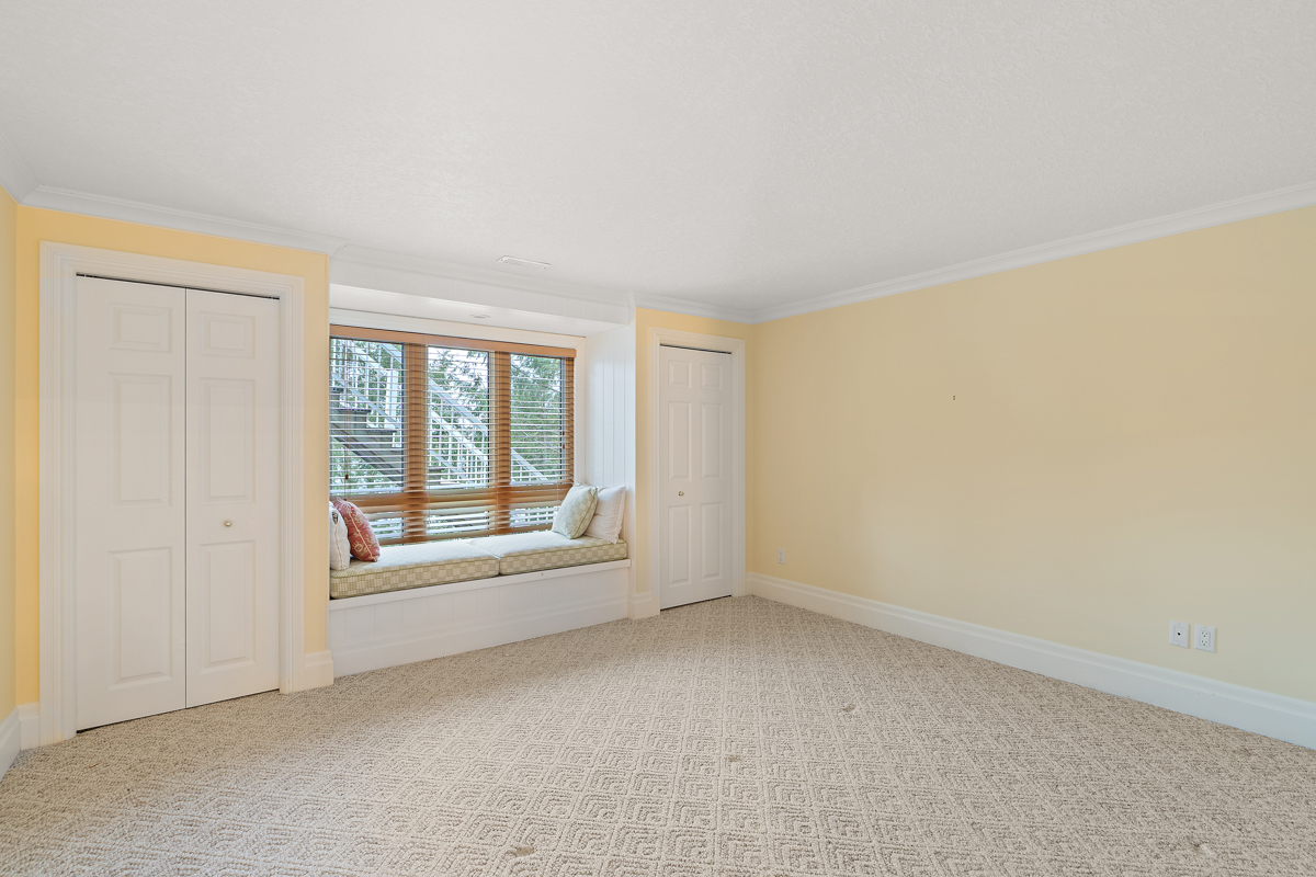 A bright, empty room with a big window. A window seat sits in front of the window, flanked by closets. The walls are yellow and the floor is carpet.