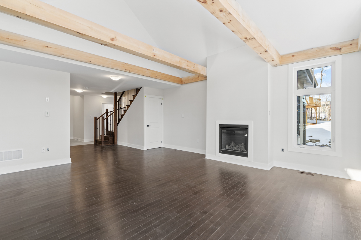 A bright, empty space with dark hardwood floors, white walls, a fireplace, and exposed beams of light wood spanning a white vaulted ceiling.