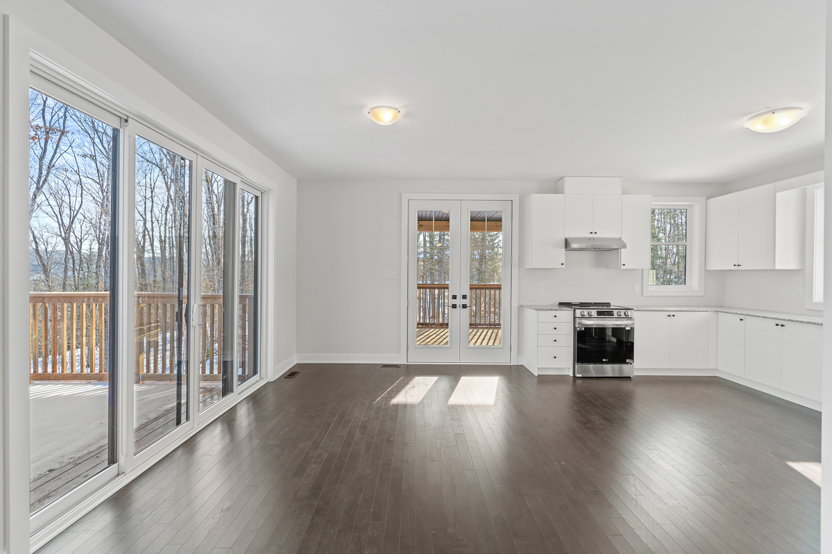 A bright kitchen area with dark hardwood floors, white walls and a white ceiling. Doors in adjacent walls lead out to different outdoor spaces. The kitchen has white cupboards and modern stainless steel appliances, and there is no furniture in the space.