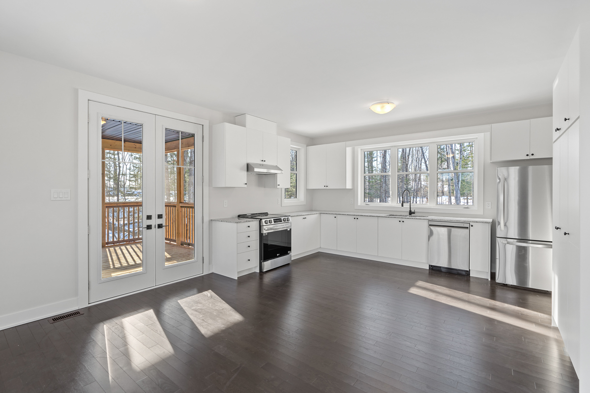 A bright kitchen area with white cupboards and modern stainless steel appliances. The area has dark hardwood floors, white walls and a white ceiling. Double doors lead out to a deck. There is no furniture in the space.