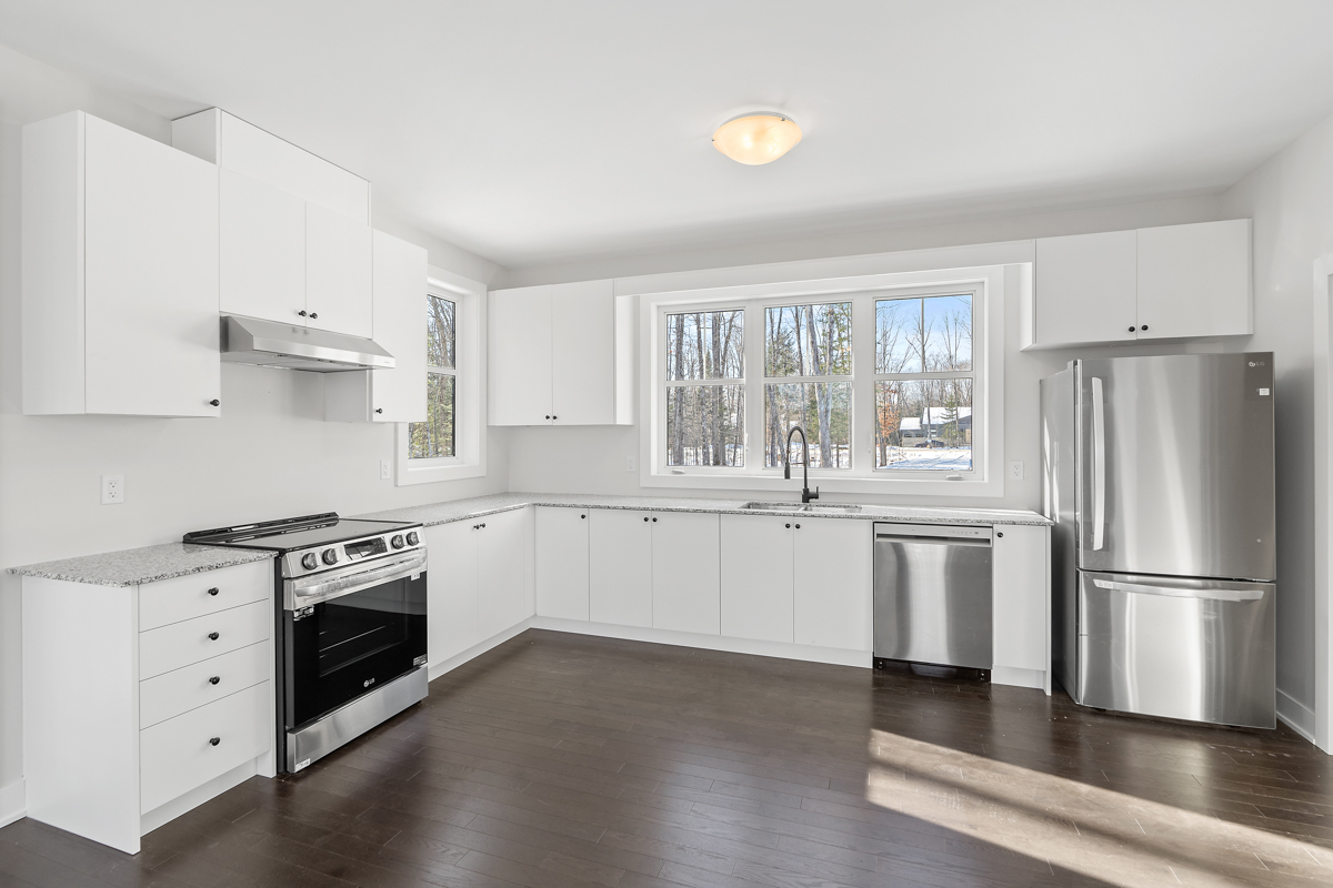 A bright kitchen area with white cupboards and modern stainless steel appliances. The area has dark hardwood floors, white walls and a white ceiling.