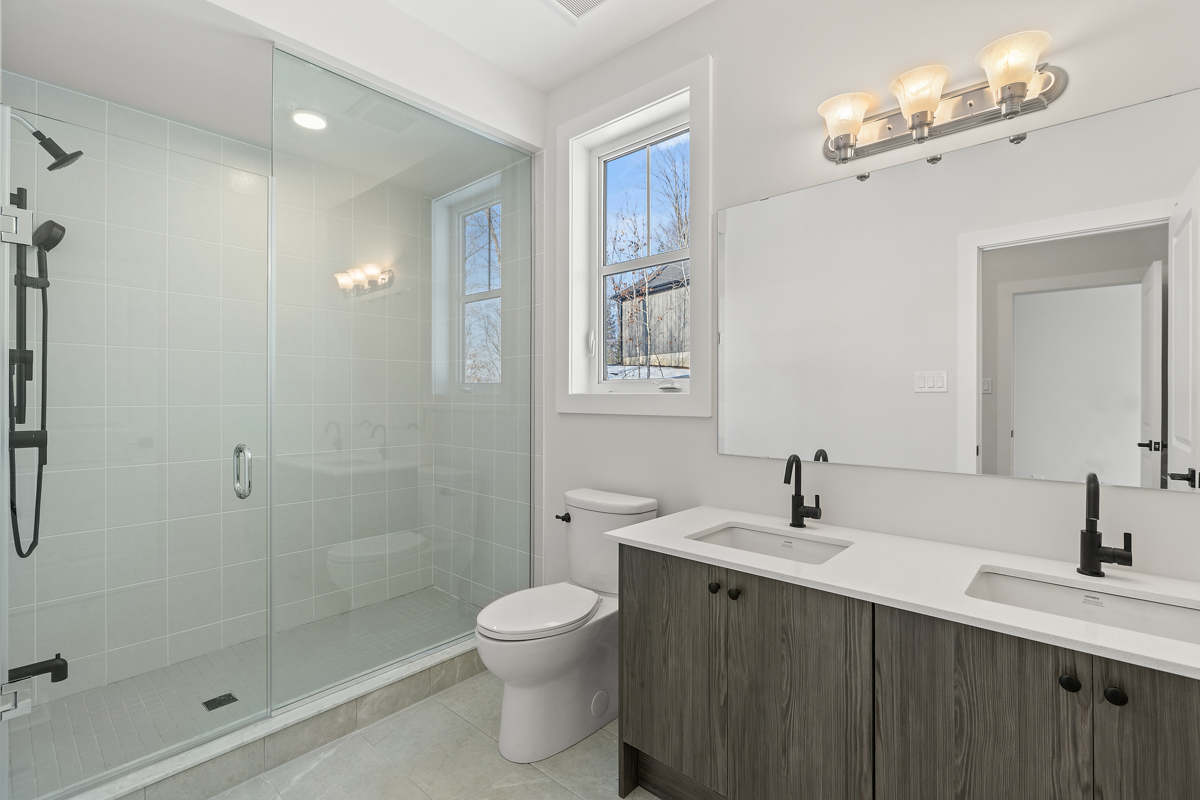 A big bathroom with a toilet, double sinks, and a walk-in shower. A small window lets in natural light.