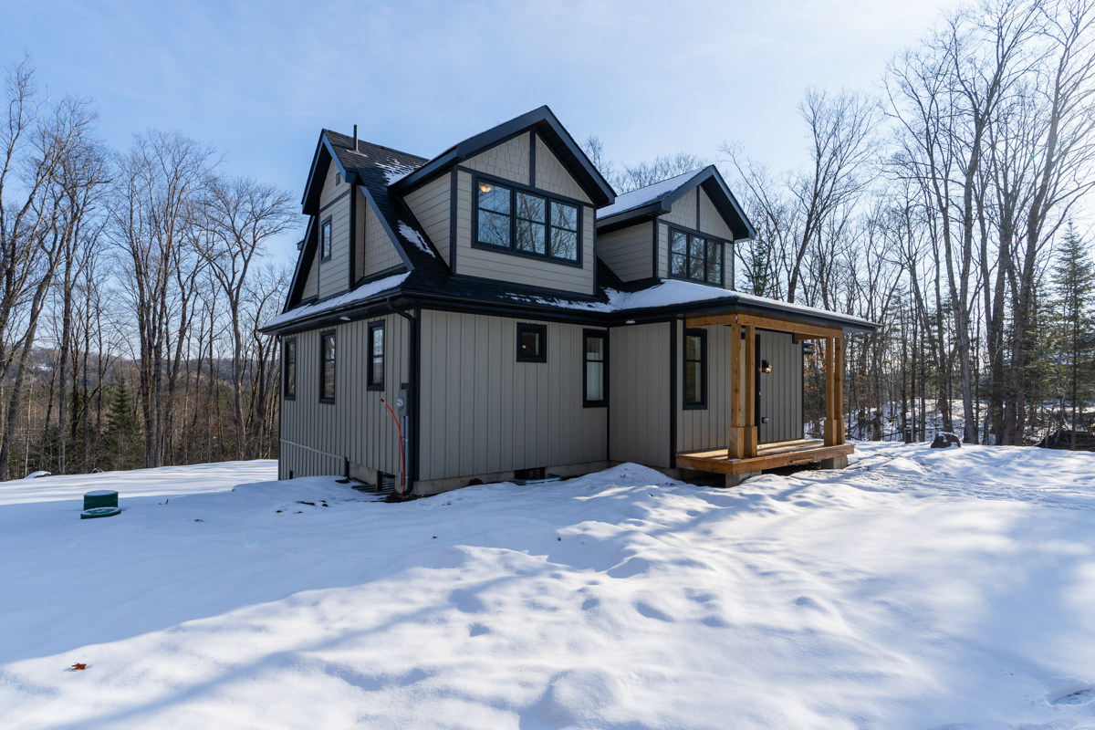 Side view of a large two-story house in a snowy, rural area. The house has a wood front porch and big windows with black trim.