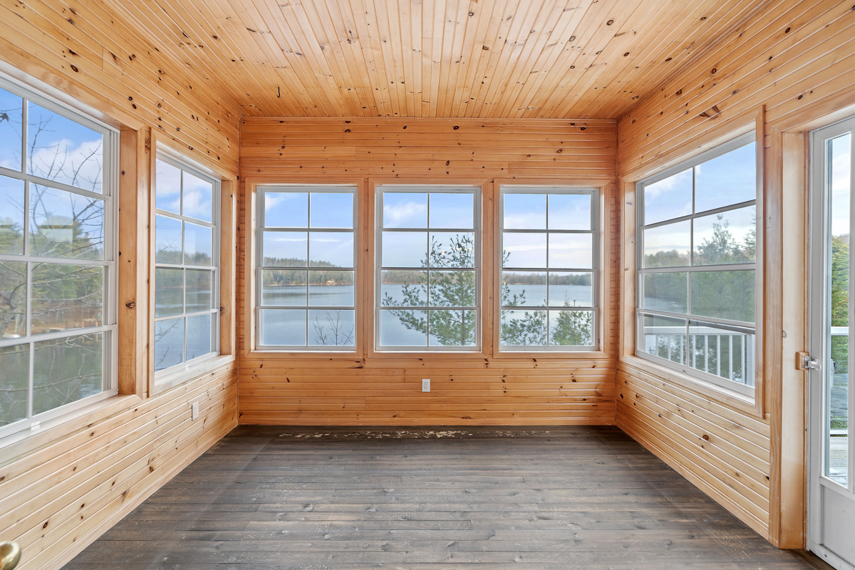 A big sunroom with lots of windows and a wood-panelled ceiling and walls, looking out over a lake.