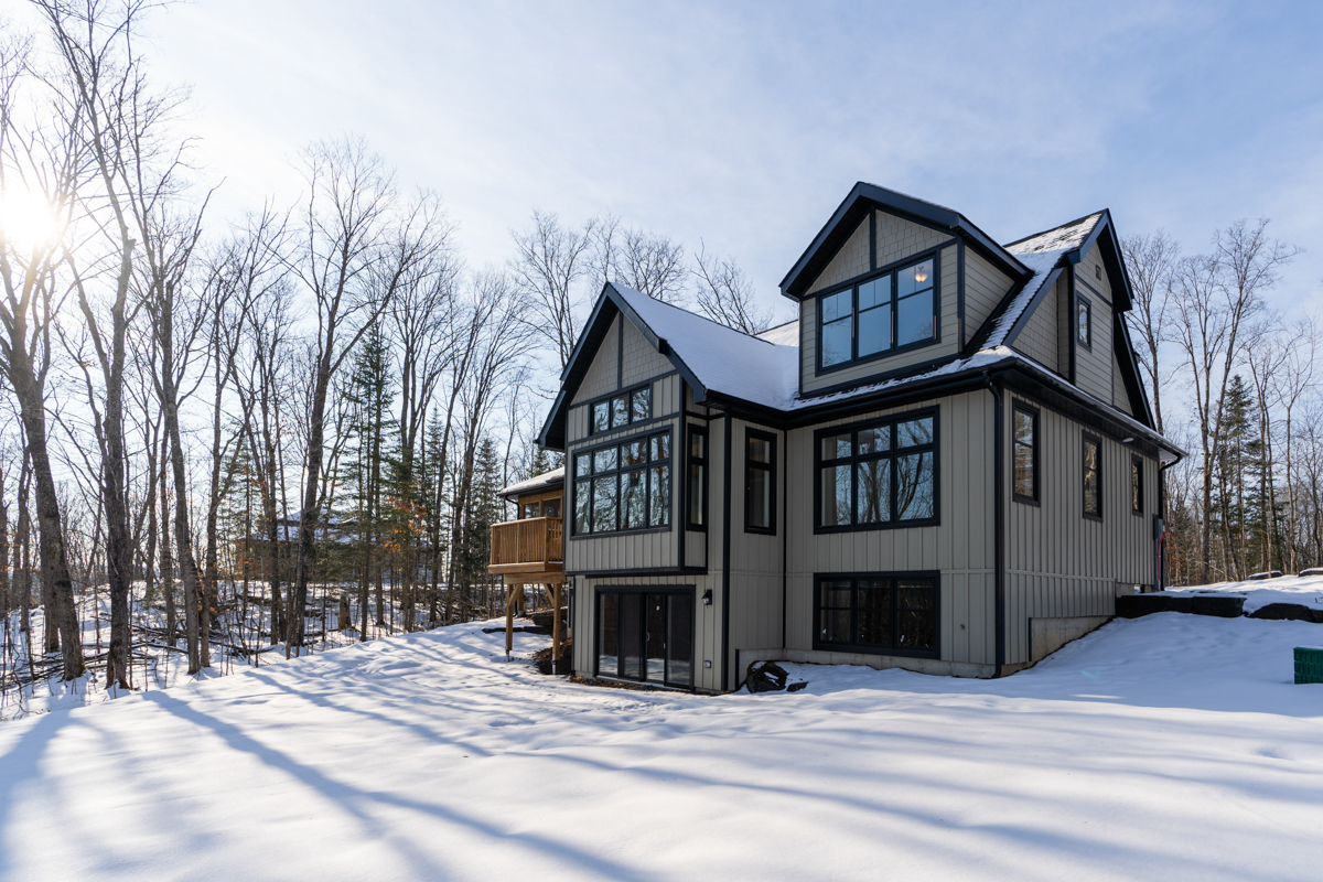 The back of a large two-story house in a clearing of a wooded, rural area. The house has big windows with black trim, and there is snow on the ground.