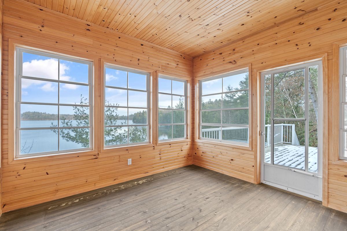 A big sunroom with lots of windows and a wood-panelled ceiling and walls, looking out over a lake.