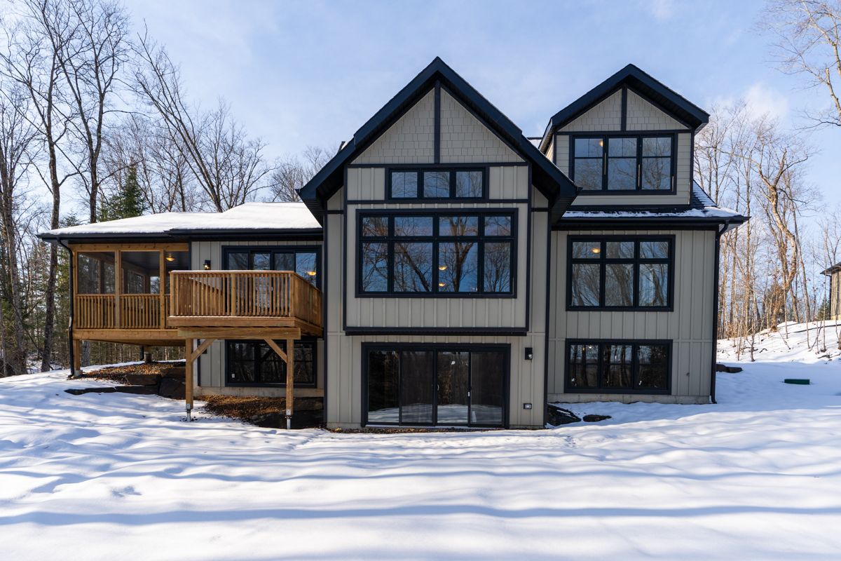 The back of a large two-story home in a rural area. The house has big windows with black trim, a raised deck, and a sunroom that extends off the back of the house. There is snow on the ground.