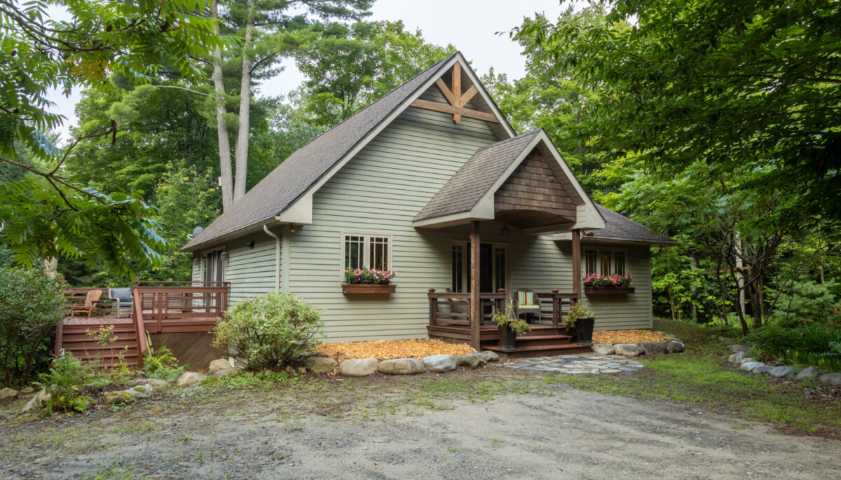 A cottage with light green siding, a slanting roof, and a big side deck sits on a dirt and gravel driveway, surrounded by green trees.