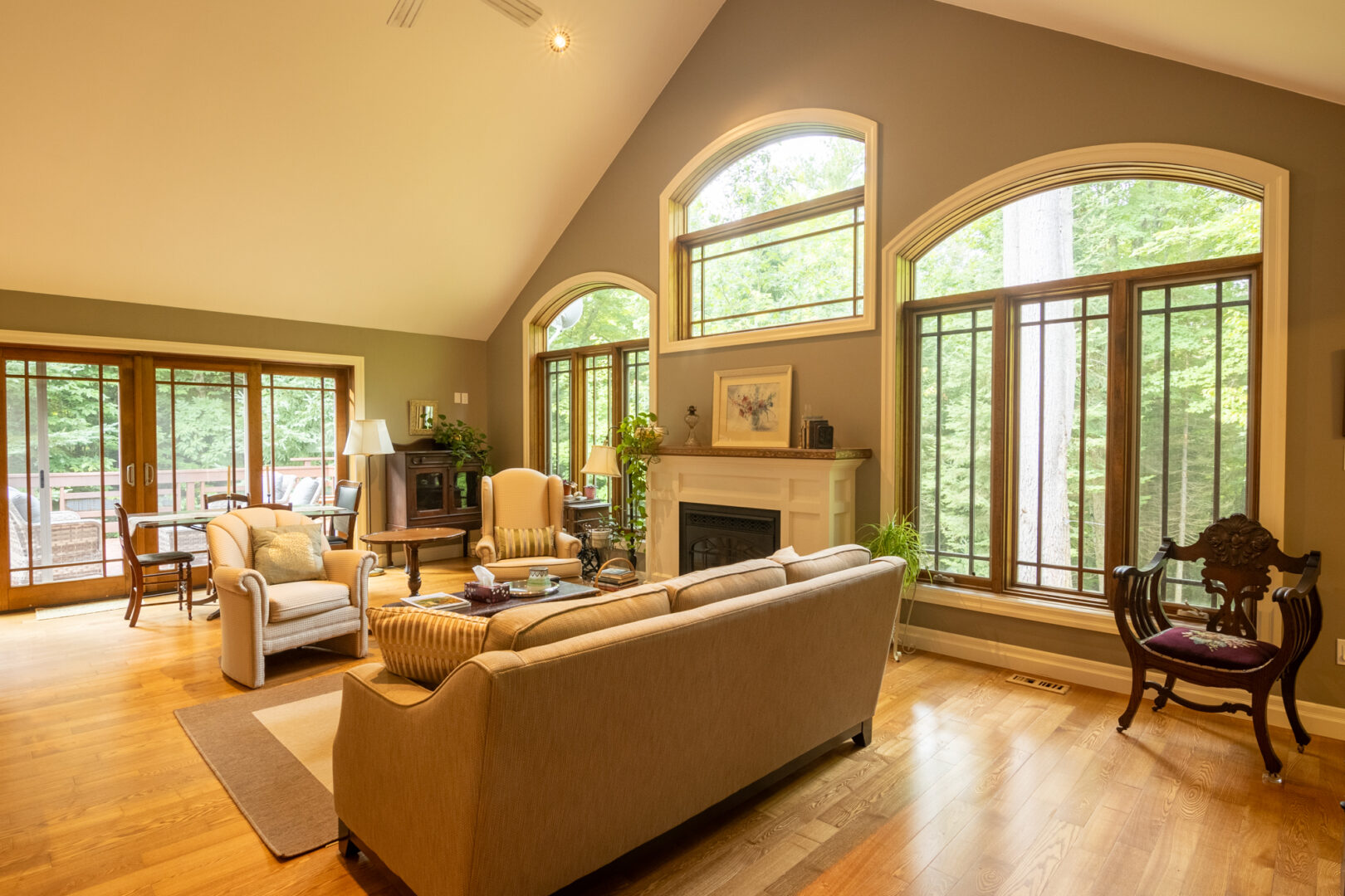 A bright living area with hardwood floors, a couch, and a high, slanting ceiling. Big windows framed in warm wood let lots of light into the space, and double glass doors with wood framing open out to an outdoor deck on the far wall.