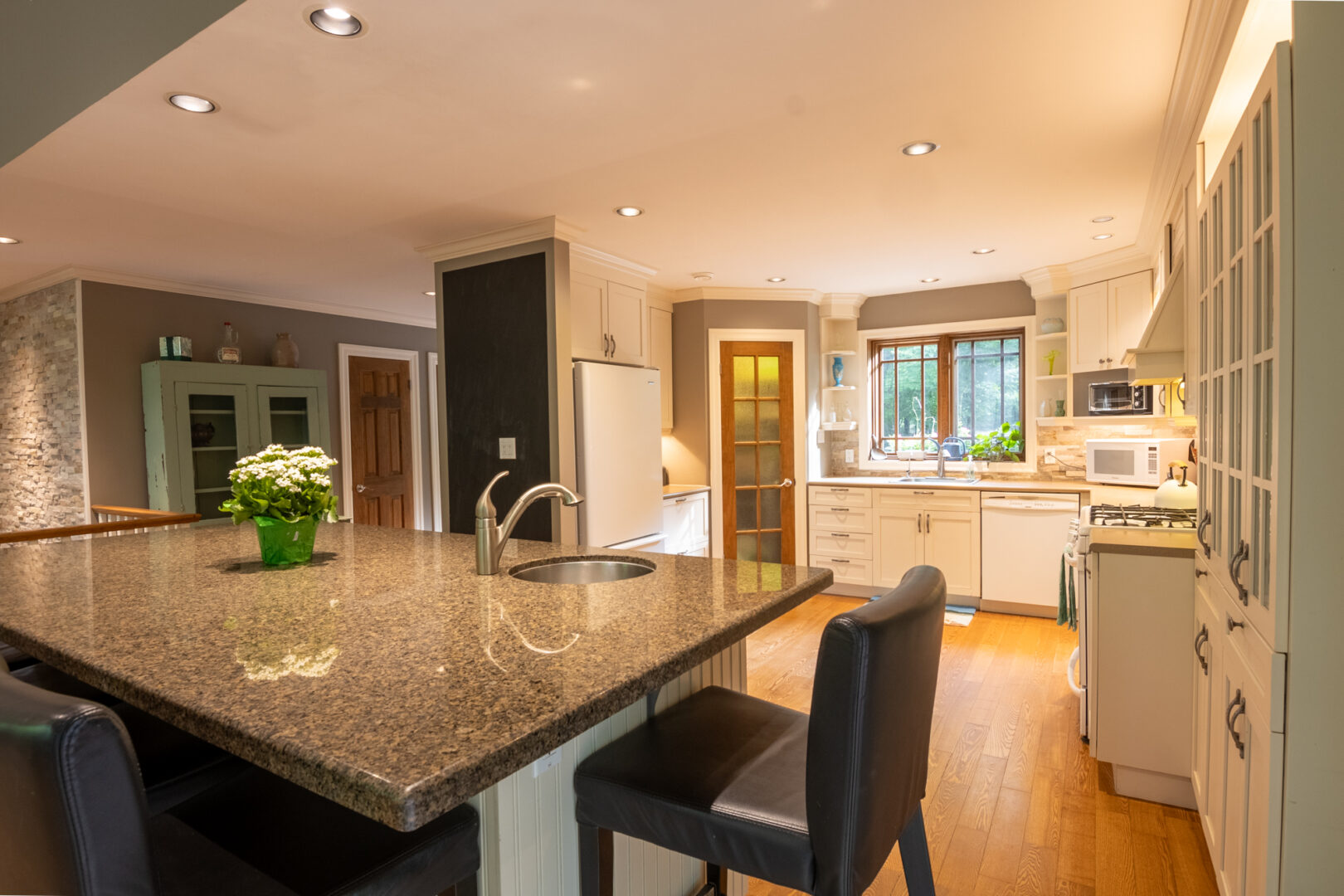 A large kitchen island with granite countertop, a sink, and barstool seating, just in front of a kitchen area. The kitchen has white cabinetry and hardwood flooring..