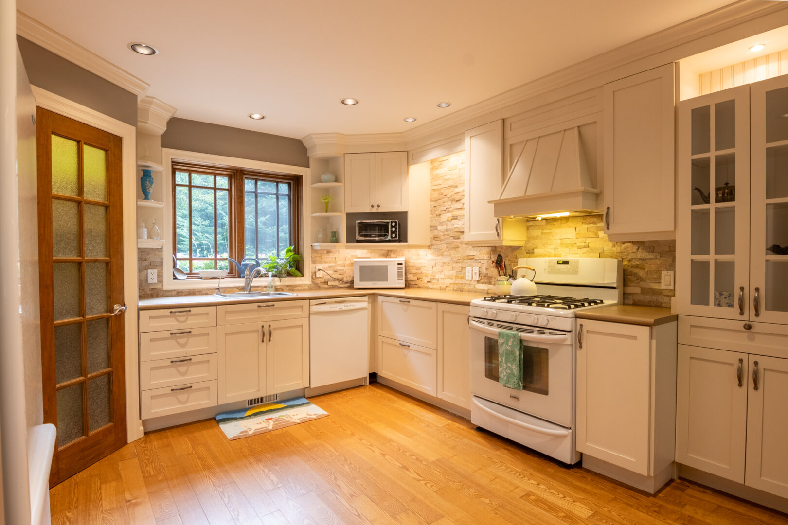 A wide kitchen space with white cabinets, hardwood flooring, and a window.
