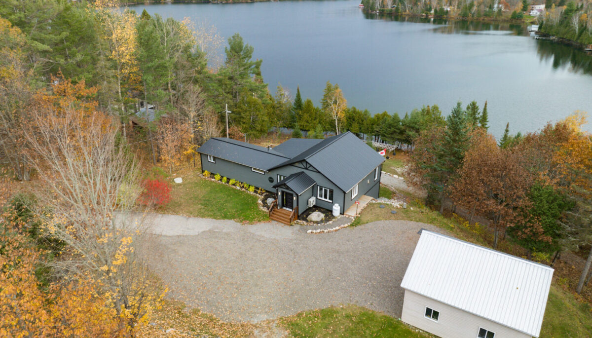 A small bungalow with a blue-gray roof and siding sits waterfront on a calm lake. The front of the house has a large paved driveway, and the home is surrounded by trees and greenery.