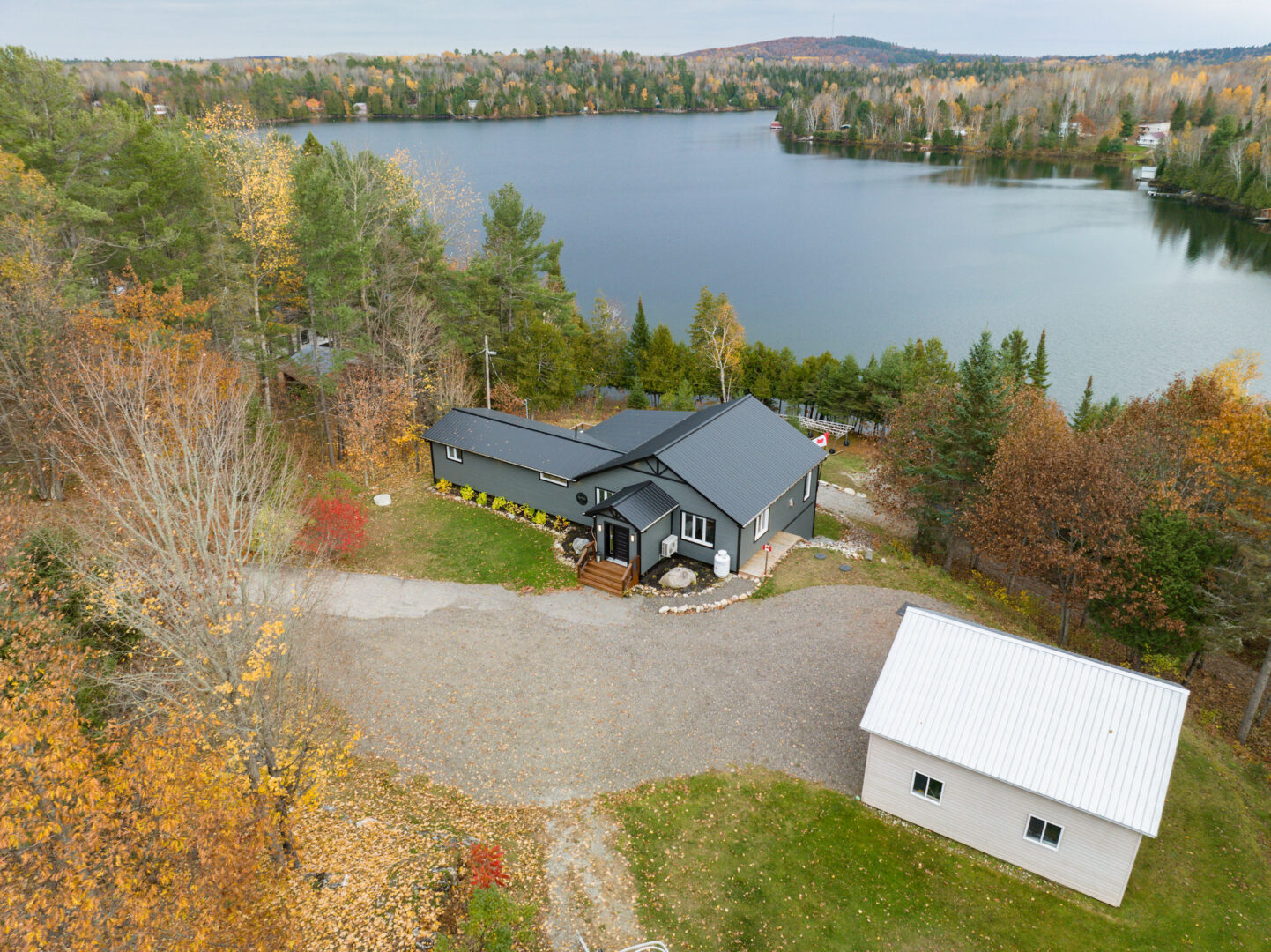 A small bungalow with a blue-gray roof and siding sits waterfront on a calm lake. The front of the house has a large paved driveway, and the home is surrounded by trees and greenery.