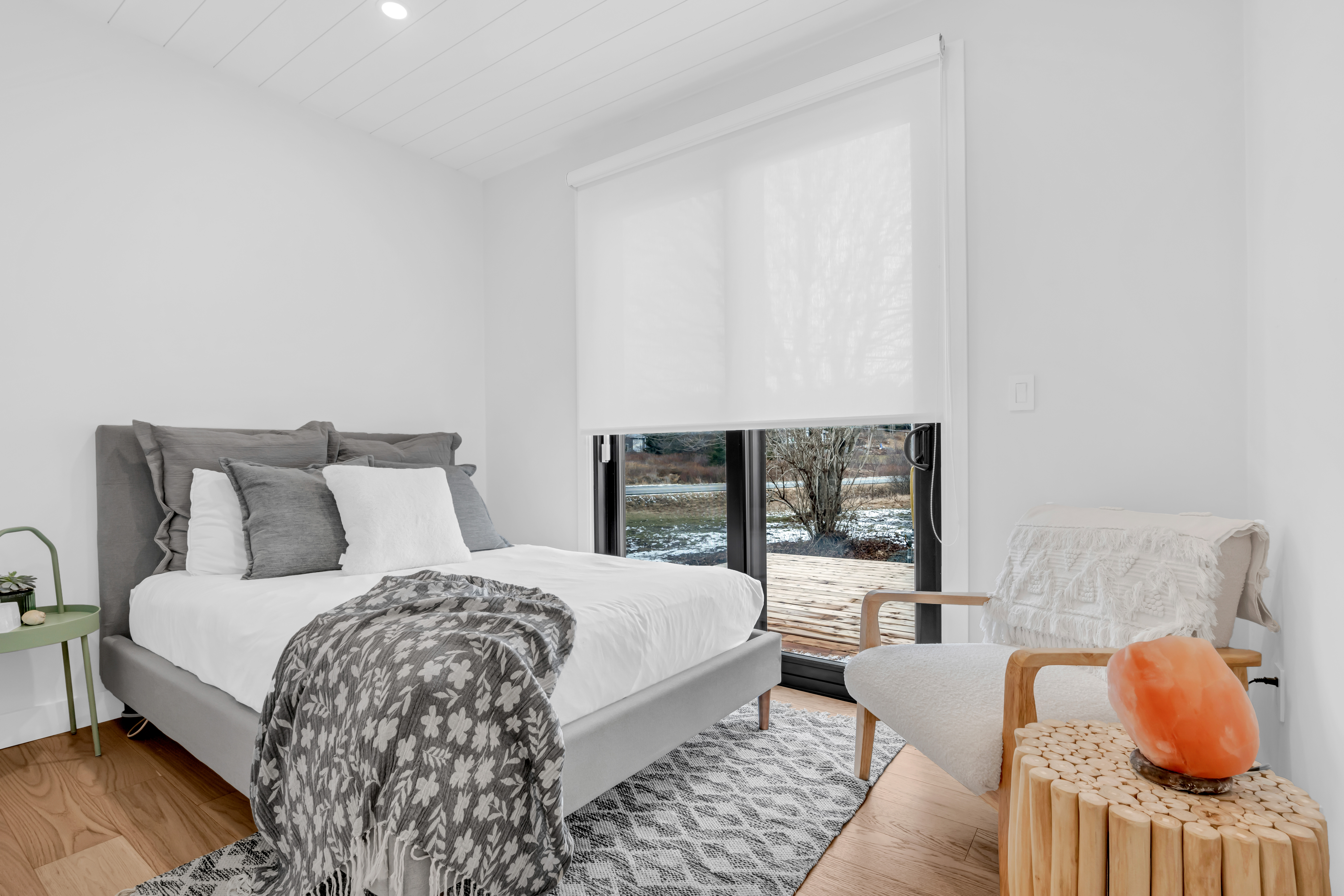 A simple bedroom with a bed, a chair, hardwood floors, and double glass doors that walk out to an outdoor deck.