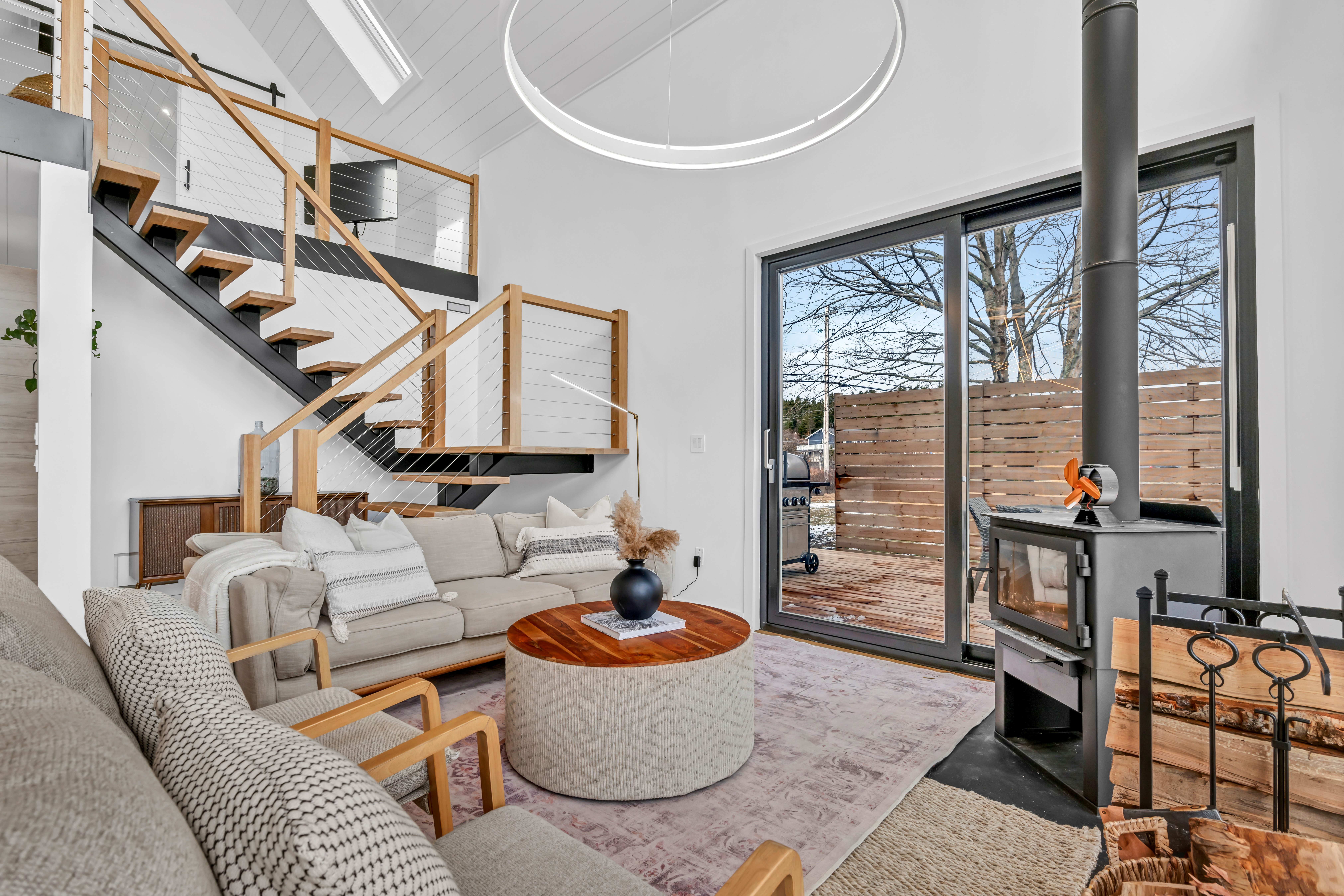 A big open-concept living area with seating and a wood stove. Floor-to-ceiling windows look out to the ocean and let in lots of natural light. A staircase leads up to a second level.