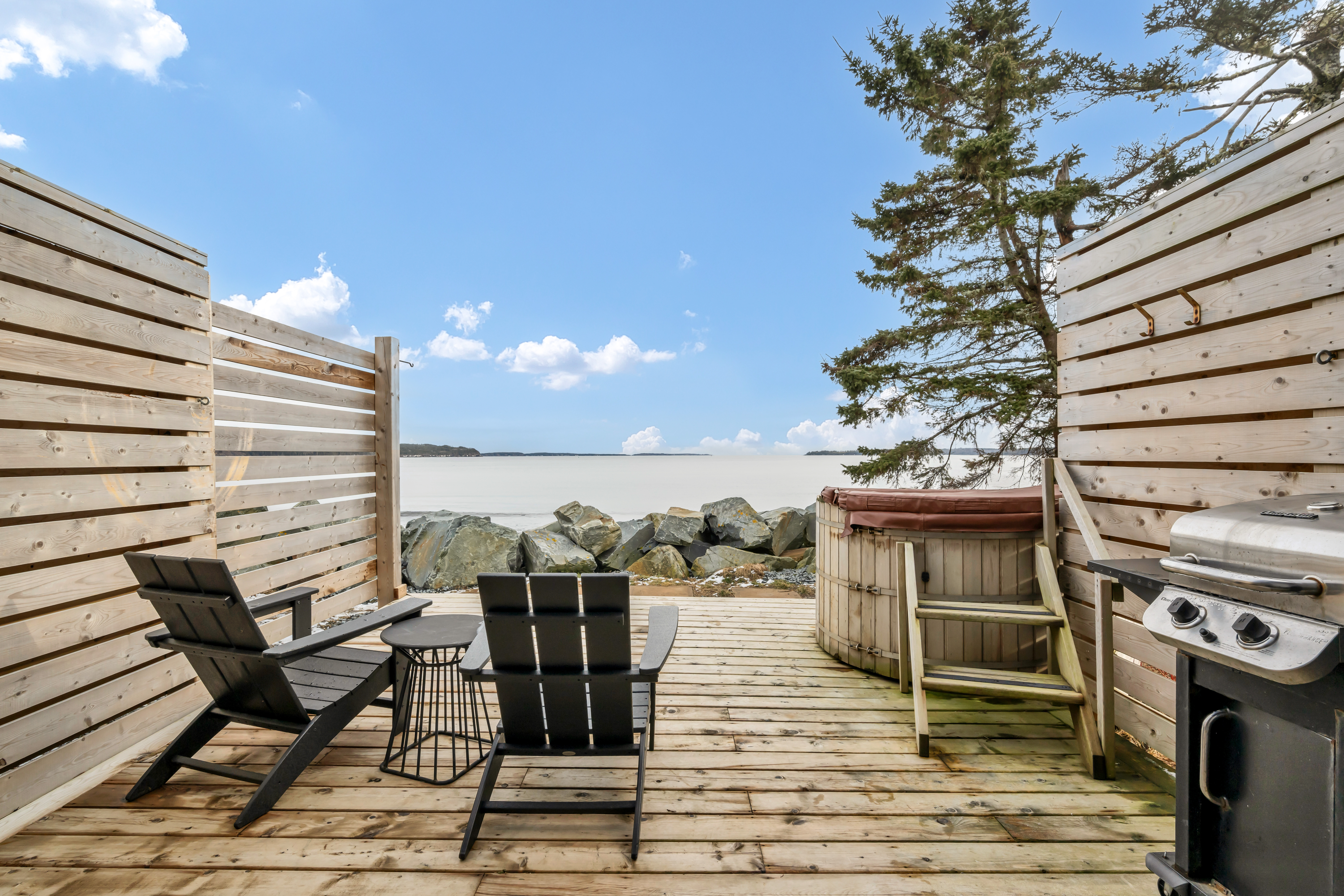 A big exterior deck off an oceanfront home that has a hot tub, a barbecue, patio furniture, and a privacy fence on either side.