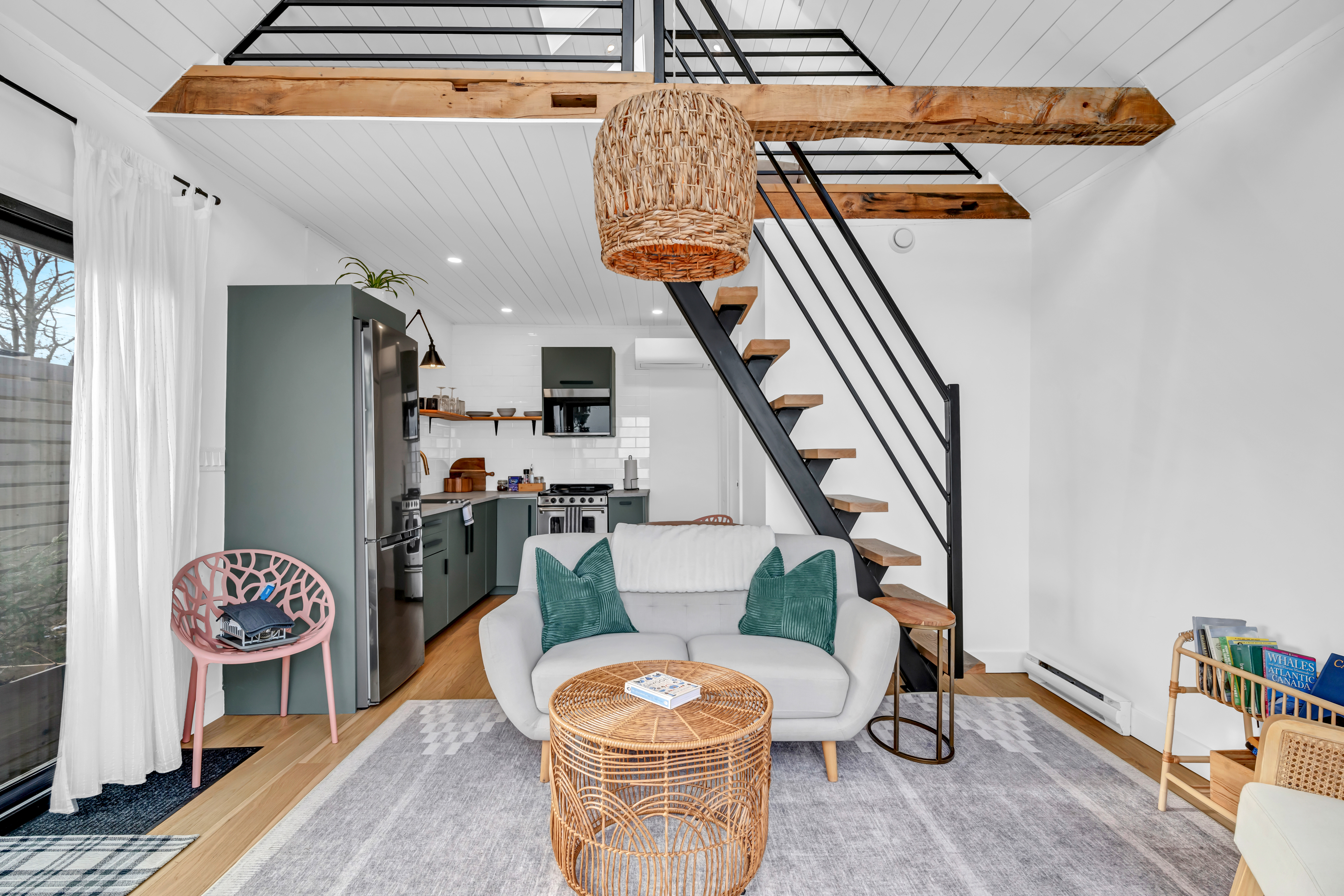 Interior of a bright, modern guest house space with an open-concept kitchen and living area. Stairs lead up to a loft level.