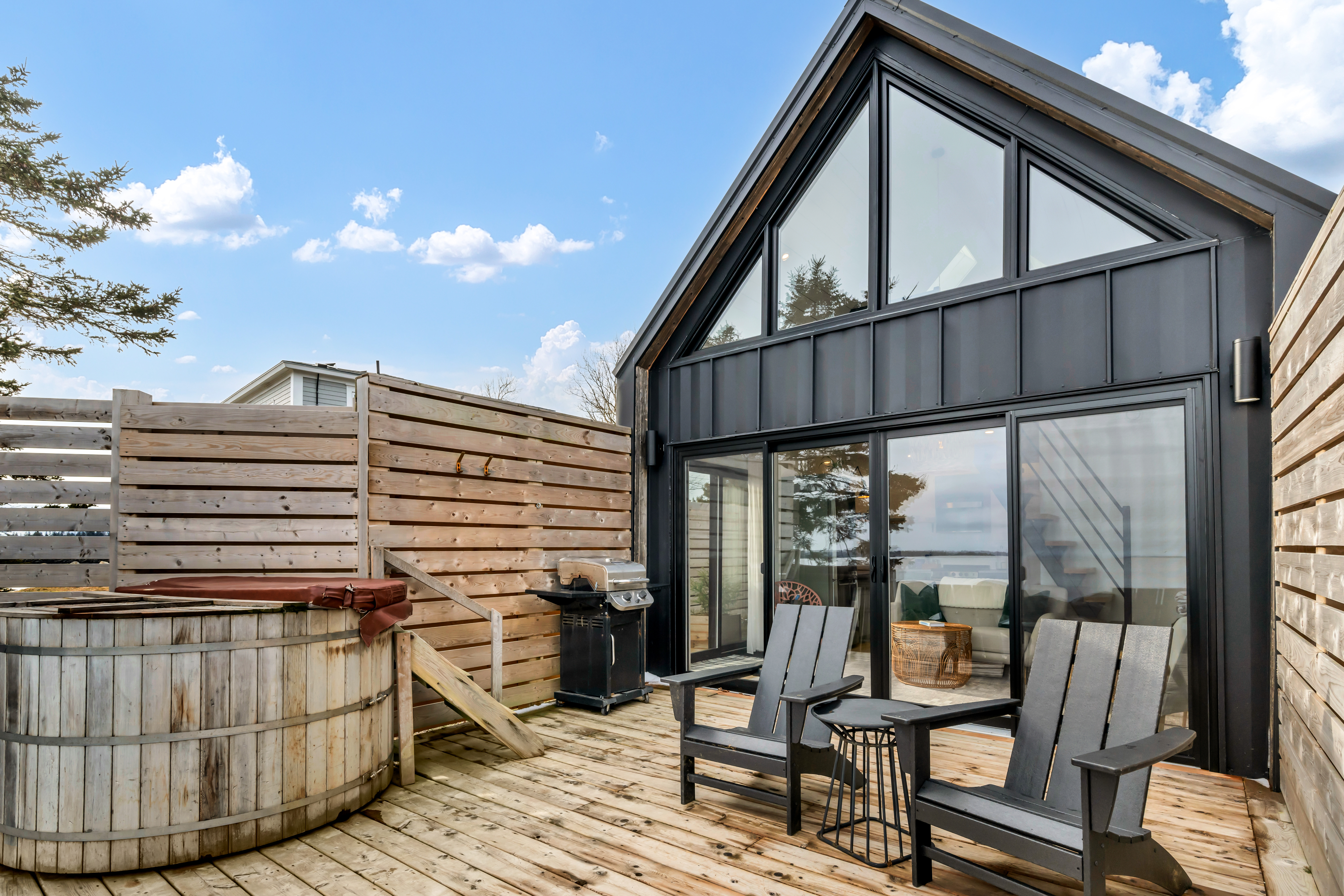 A small oceanfront guest house with large windows and a big exterior deck that has a hot tub, a barbecue, patio furniture, and a privacy fence on either side.