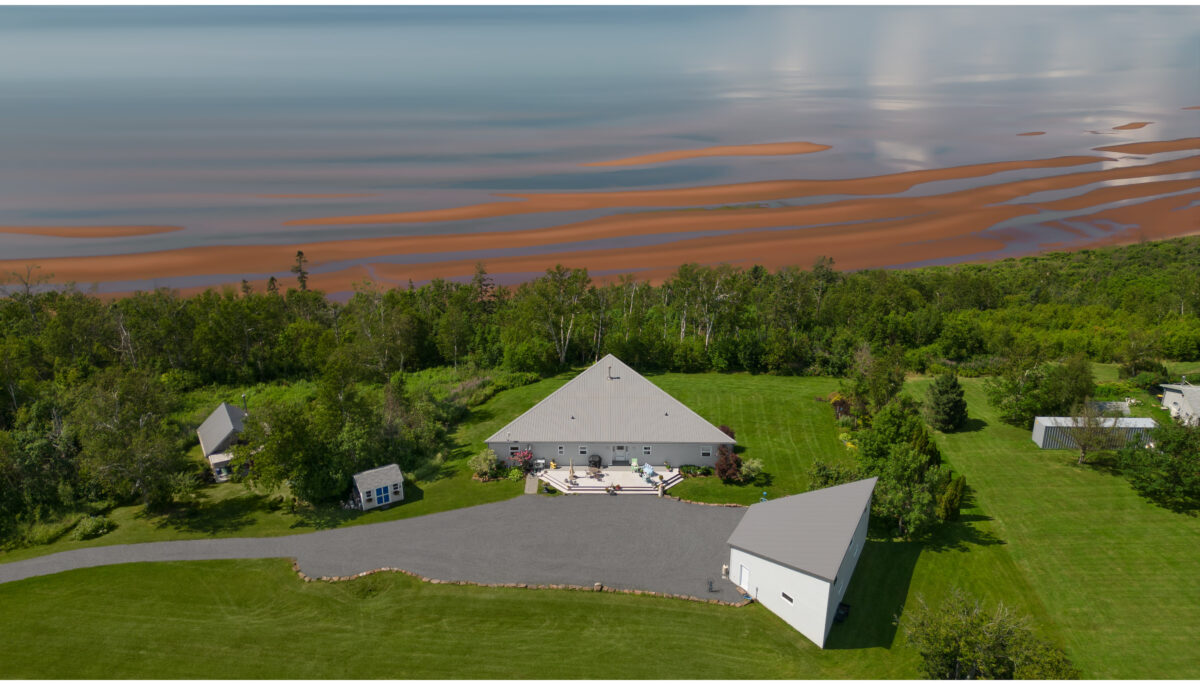 A large waterfront Prince Edward Island home surrounded by green trees, looking out to a red sand beach and a big stretch of water.