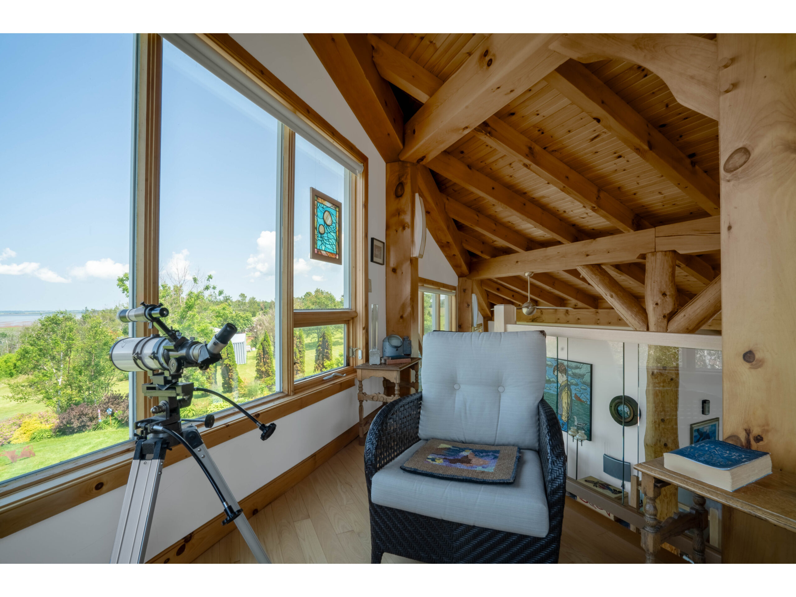 A loft space with an arm chair and a telescope in front of big windows, looking out toward green trees and water.