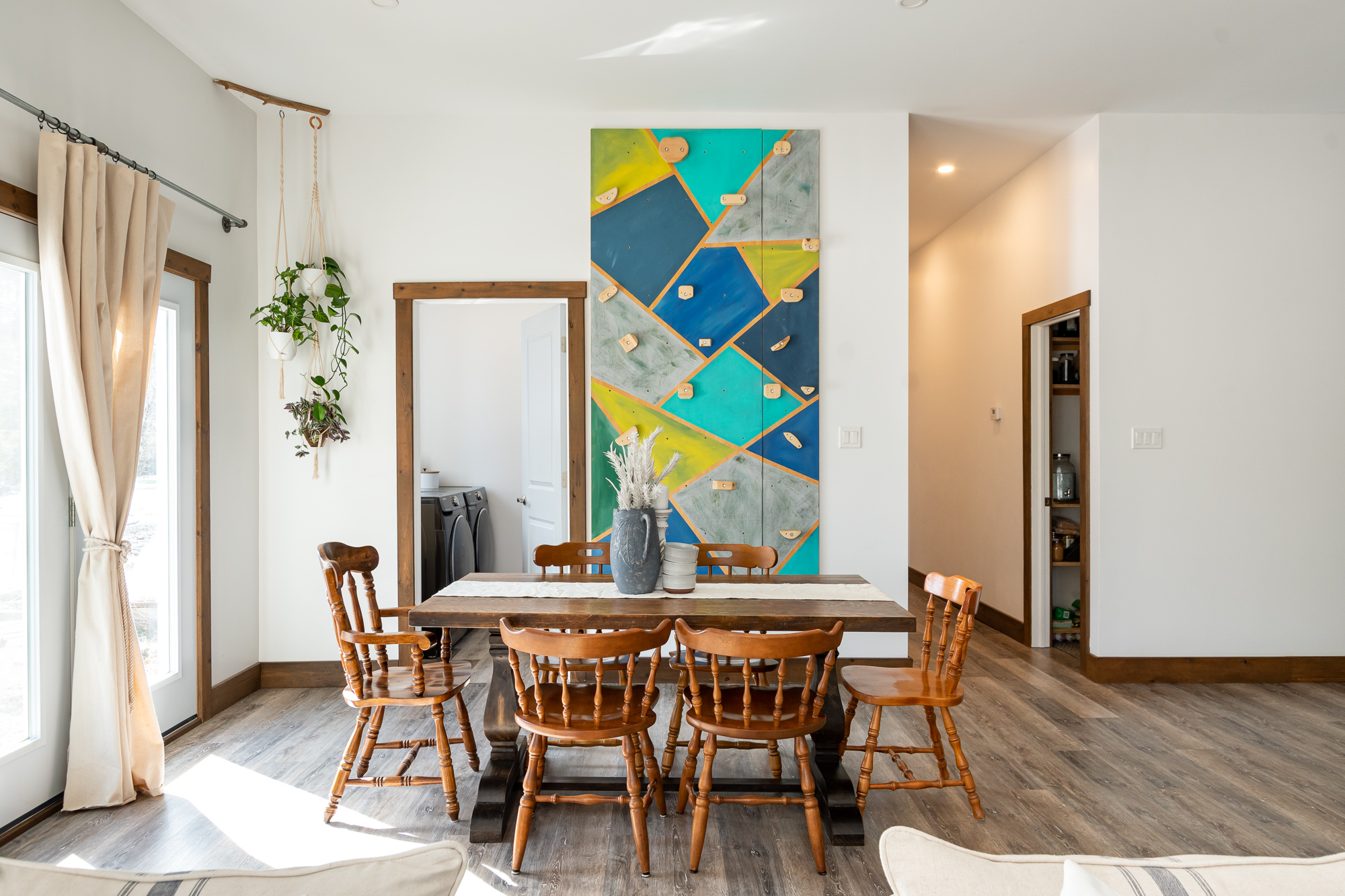 A brown dining table surrounded by chairs. A mirror and artwork that resembles a climbing wall sit on the wall behind