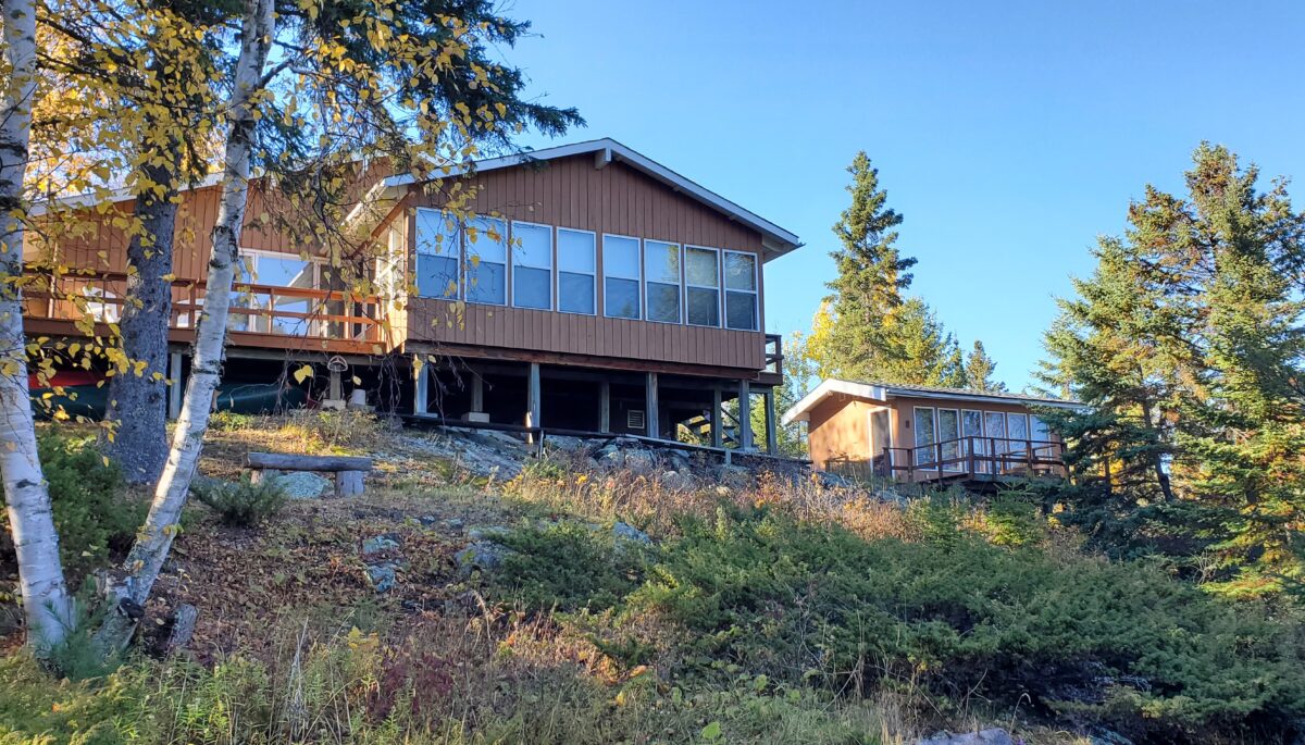 A lakefront cottage with lots of windows sits on a high slope, surrounded by trees.