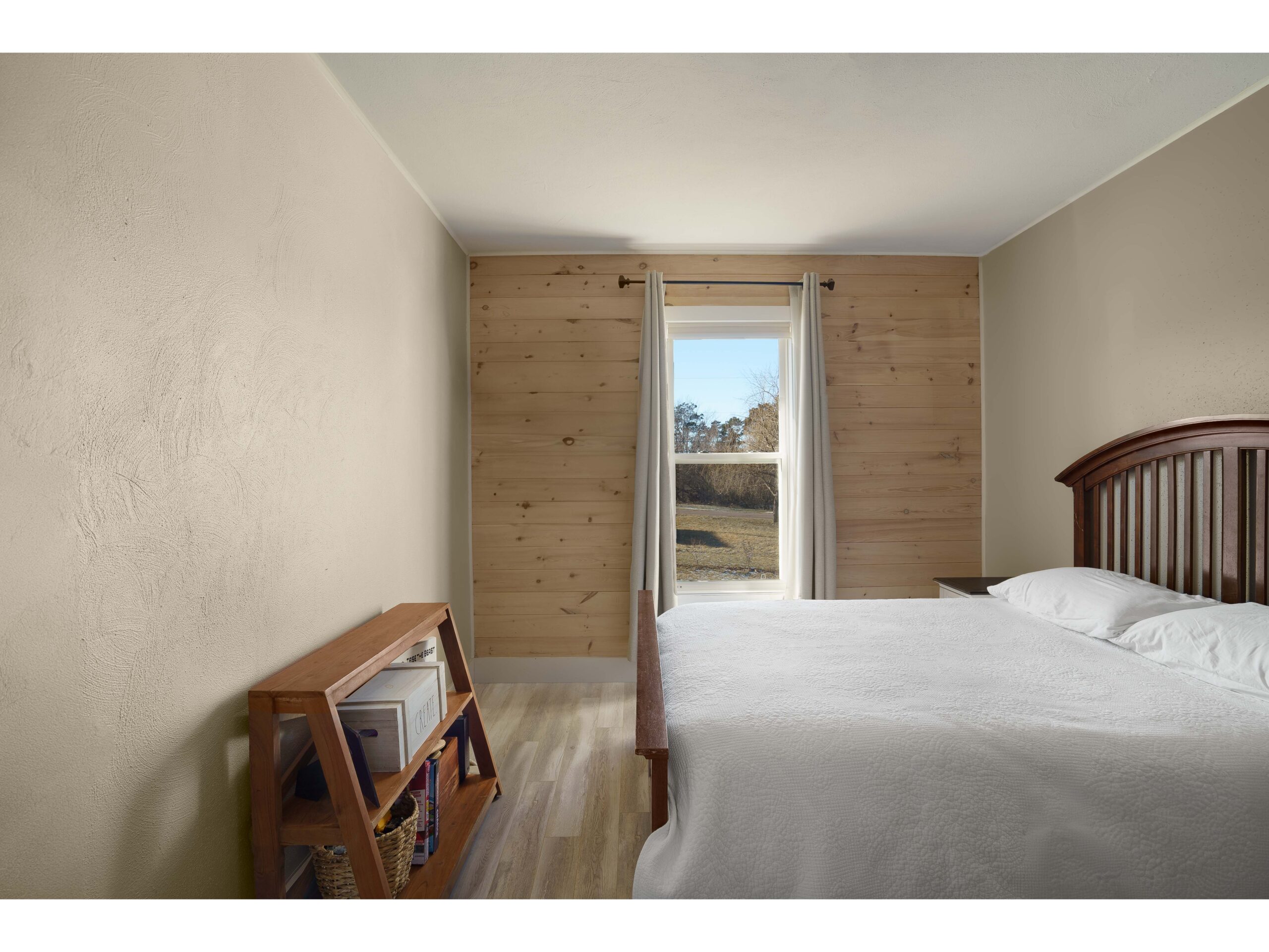 A small bedroom with a big bed, a wood-panelled wall with a window, and a small, short bookshelf.