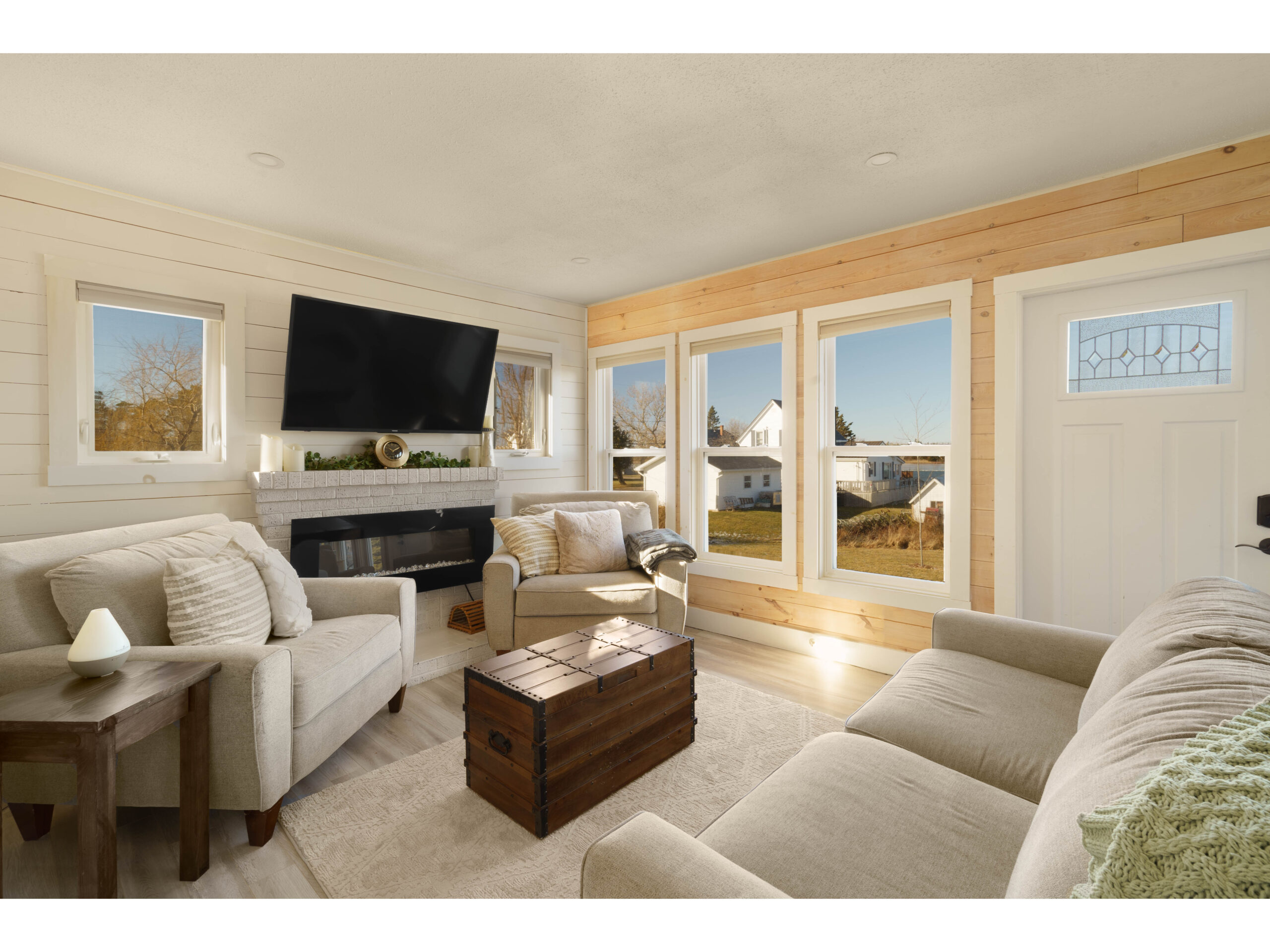 A bright but cozy living space just inside a front door, with big windows, a couch, two chairs, a television, and wood-panelled walls.
