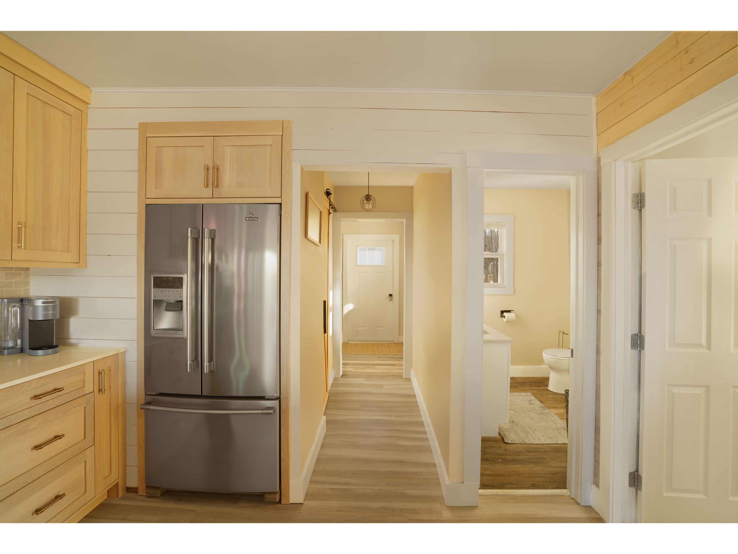 A hallway framed by a kitchen with a stainless steel fridge on one side and a bathroom on the other side. The hallway leads toward a door to outside.