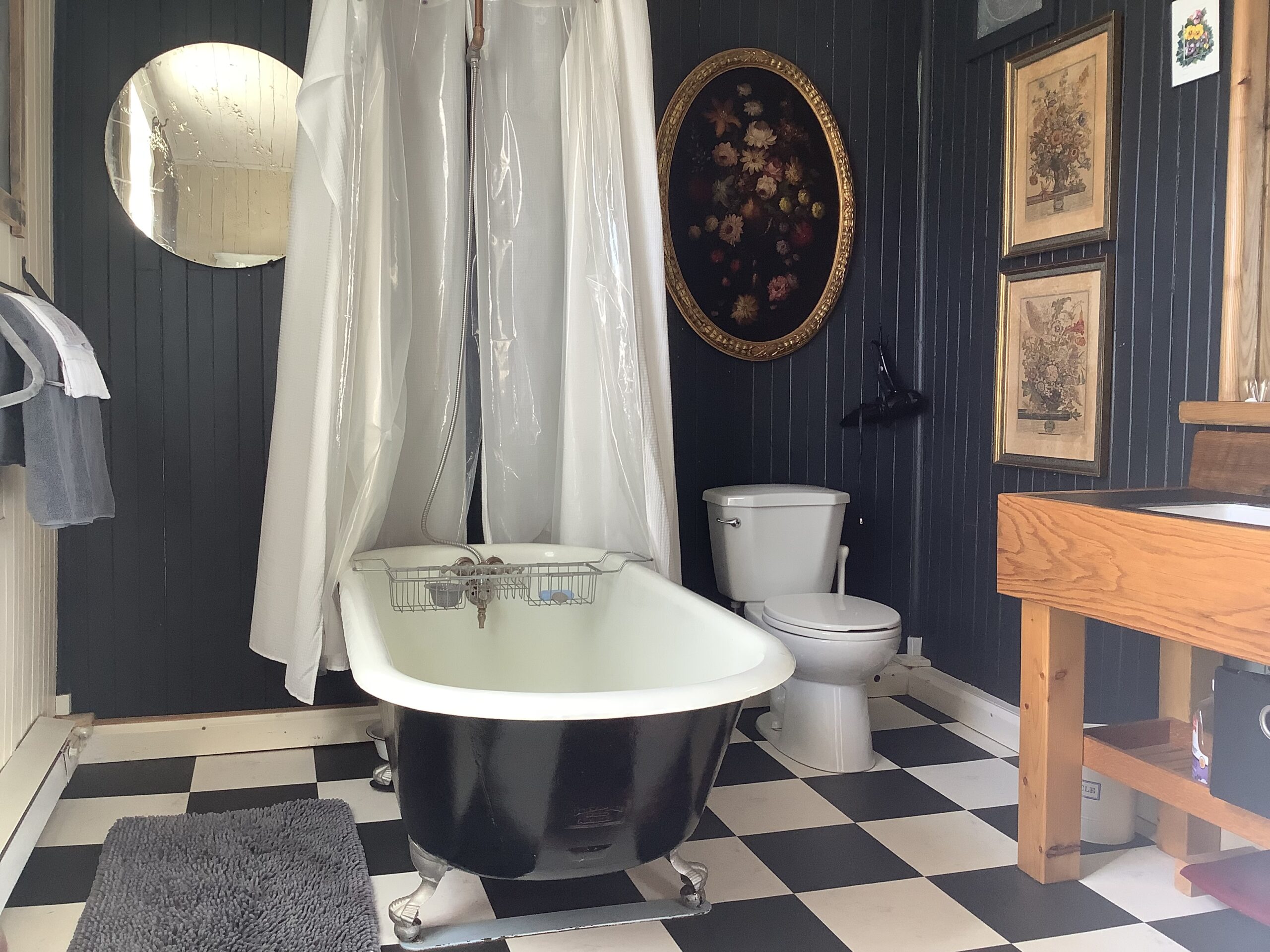 A bathroom with black-and-white checkerboard flooring, black-painted walls, a toilet, a sink, and a black clawfoot tub with a shower head and curtain attached.