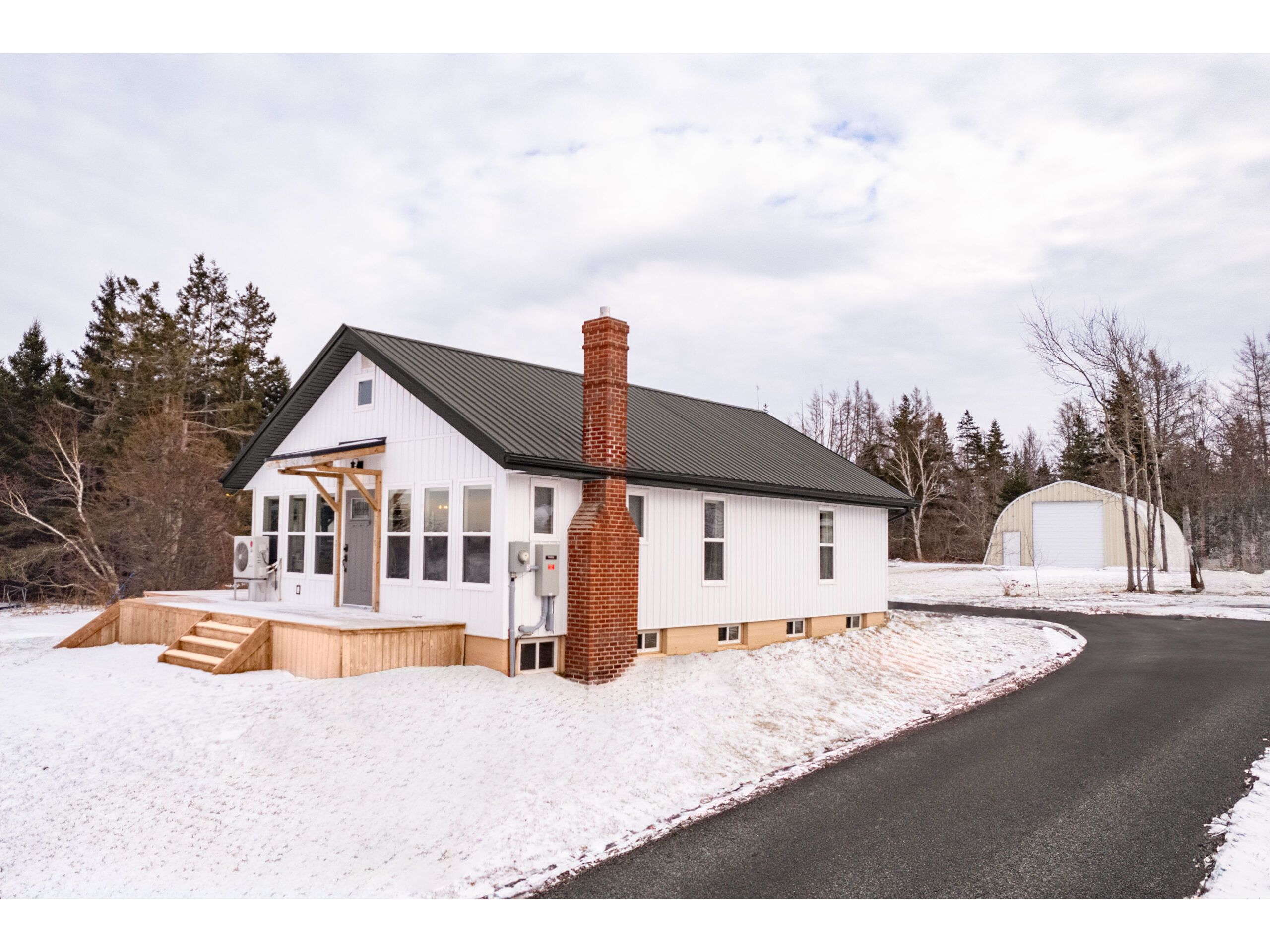A small home with a white exterior, black trim, a big wooden deck, and lots of windows, sitting on a snow-covered lot.