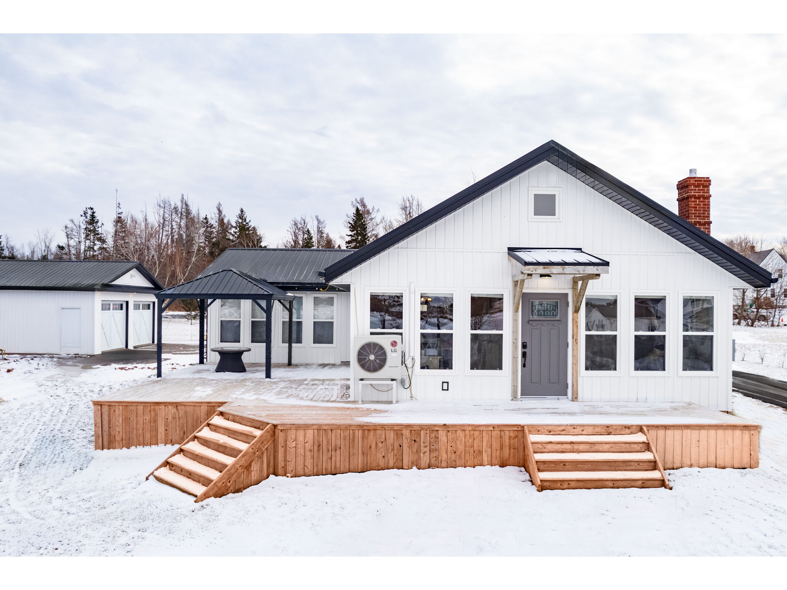 A small home with a white exterior, black trim, a big wooden deck, and lots of windows, sitting on a snow-covered lot. A detached double garage, also white with black trim, sits behind the house.