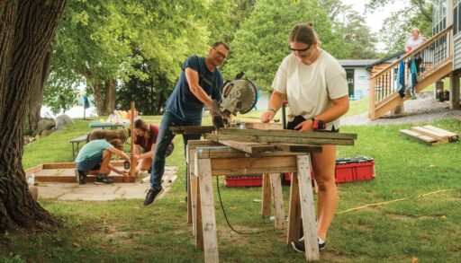 A girl and her dad use an electric saw together on a stack of lumber