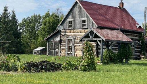 A farmhouse sits in a green field, with trees beyond.