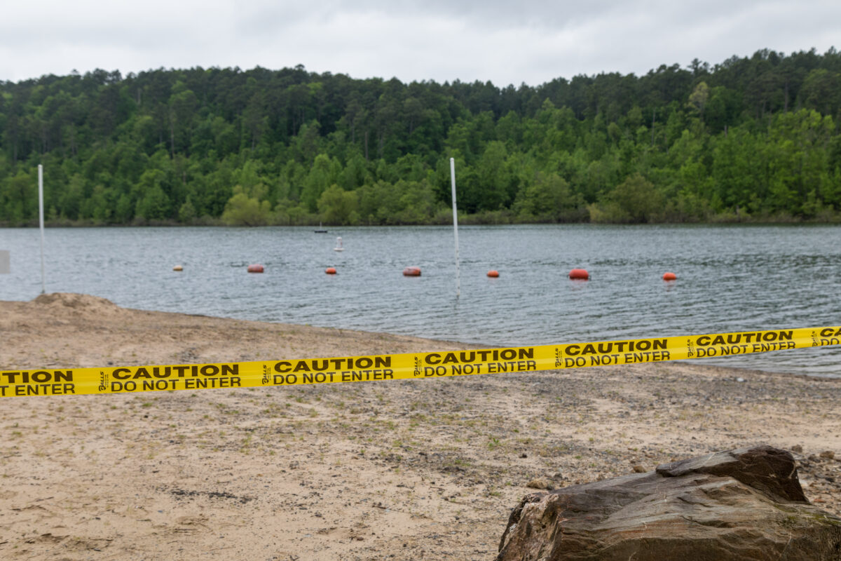 Caution tape restricts beach access in front of a lake.