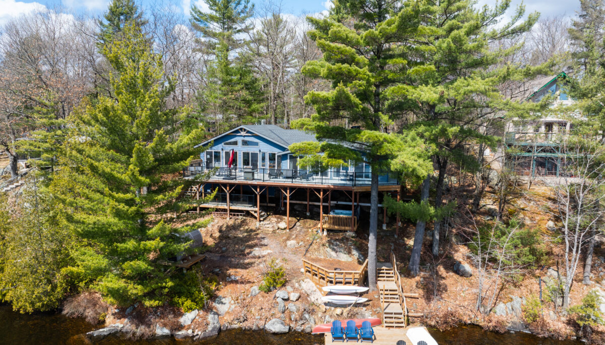 Aerial view of a blue cottage on tall stilts. Green trees surround the cottage, and brown grass on a hill leads down to a dock