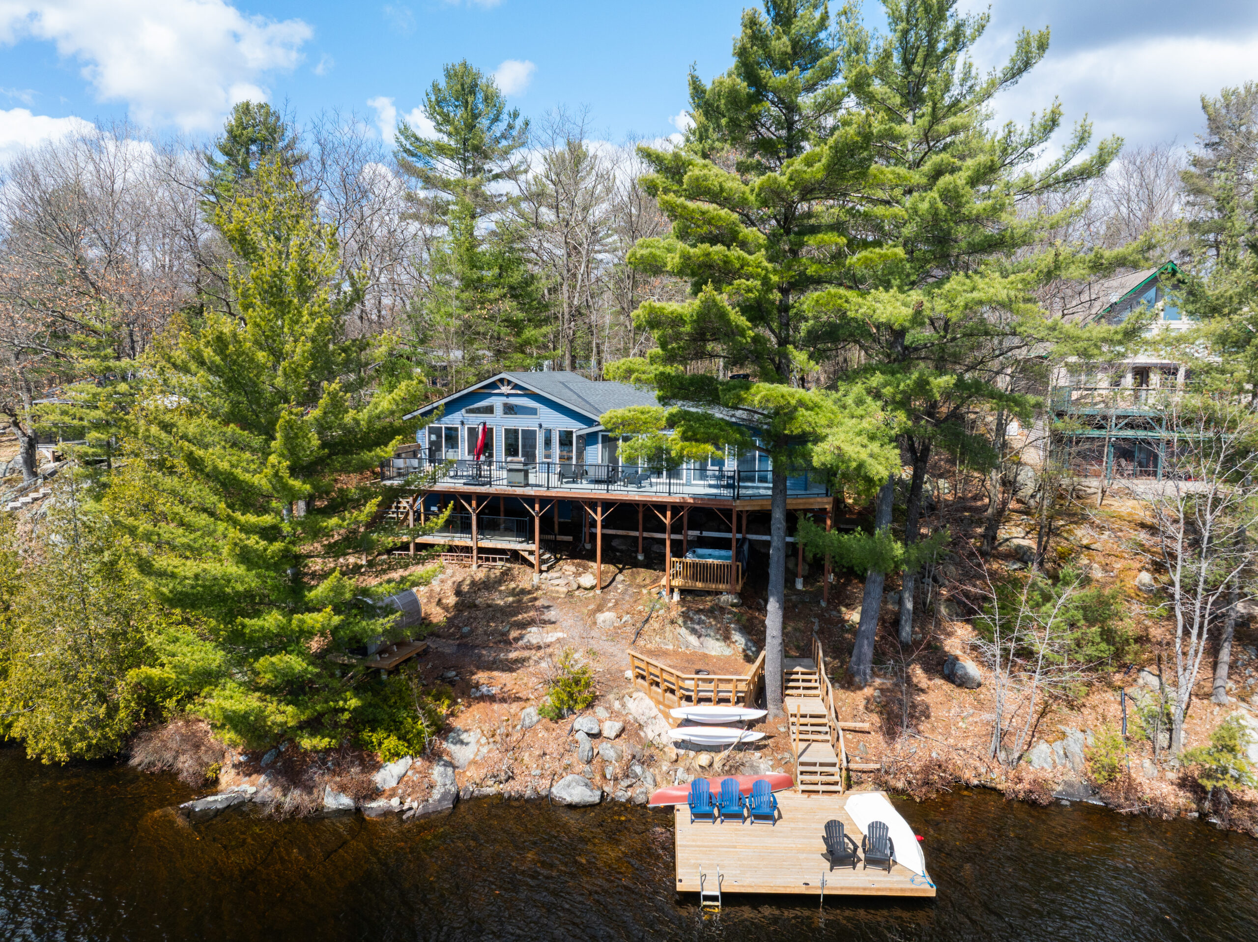 Aerial view of a blue cottage on tall stilts. Green trees surround the cottage, and brown grass on a hill leads down to a dock