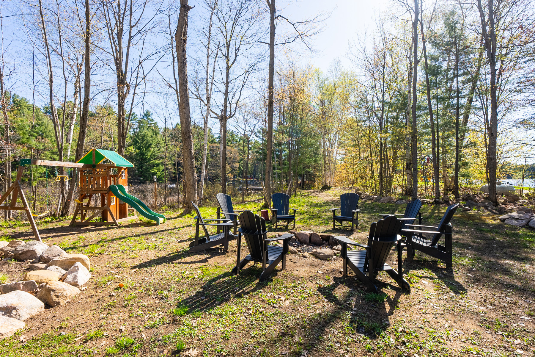 A fire pit surrounded by black Muskoka chairs, and a children's play structure with a slide, on a big, even grassy lot.