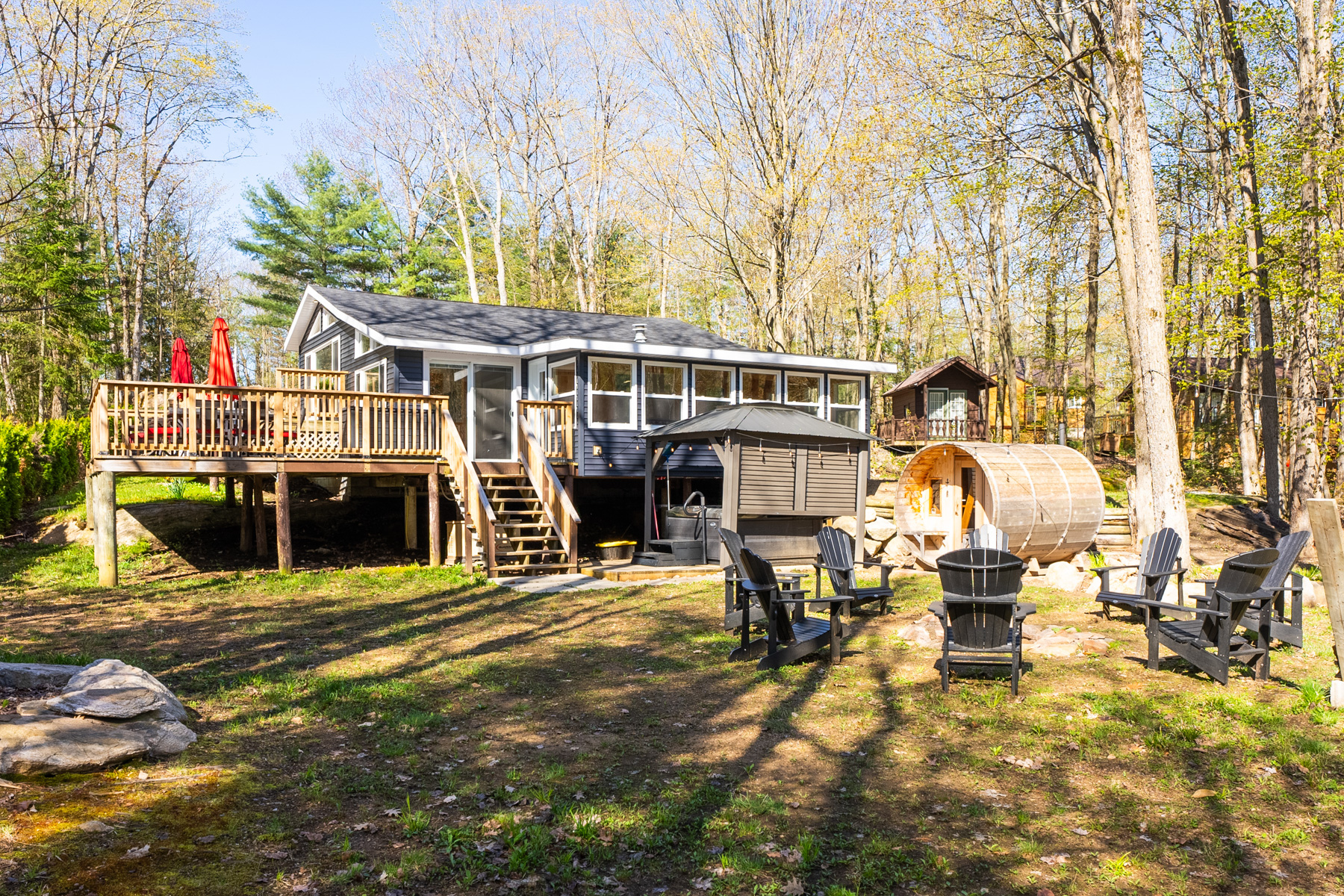 A big cottage sits on a grassy, even plot of land with lots of trees. There is a covered hot tub area, a wooden barrel sauna, a fire pit surrounded by black Muskoka chairs, and a children's play structure with a slide.
